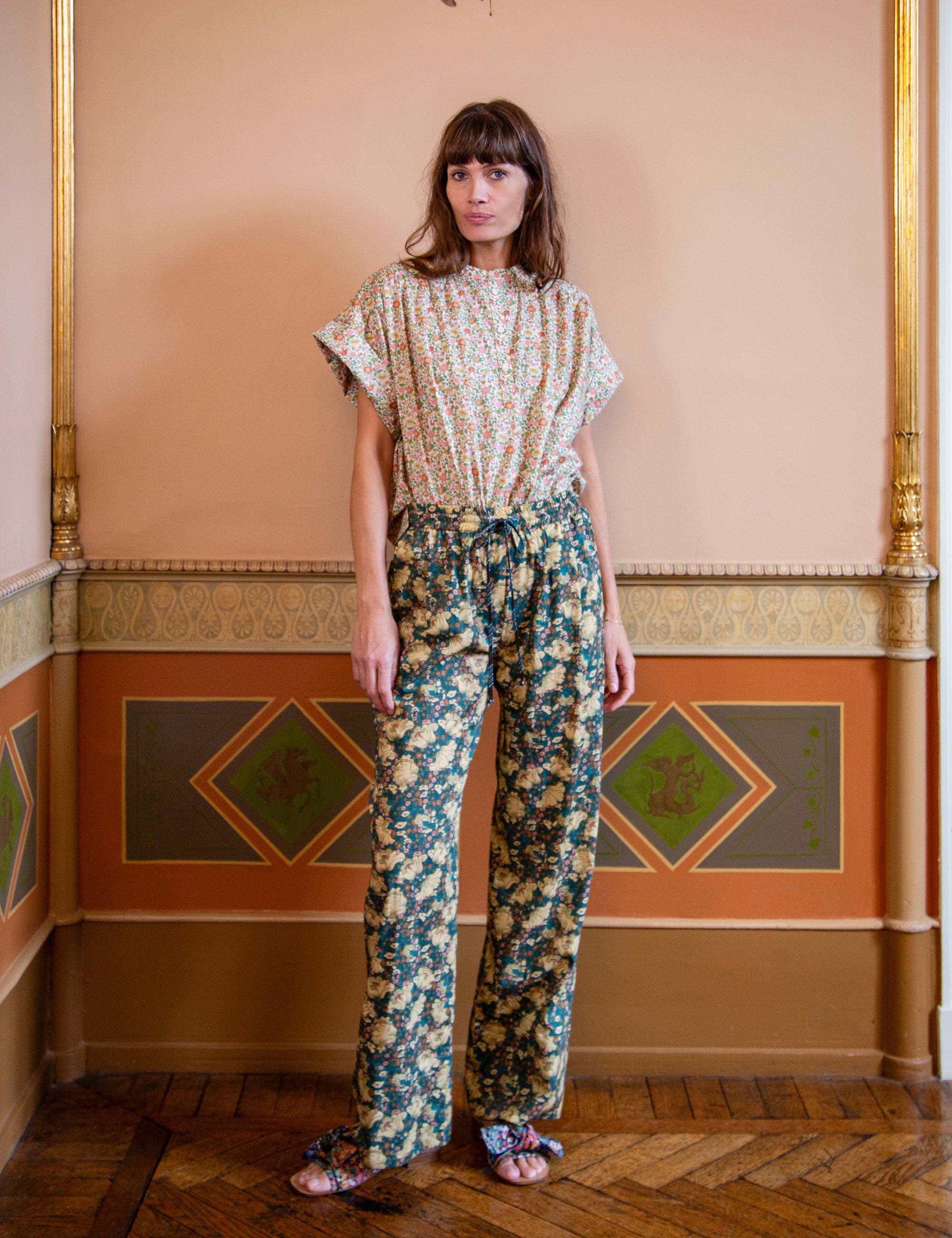Woman wearing a short sleeve floral patterned top and floral pants in an ornate room.