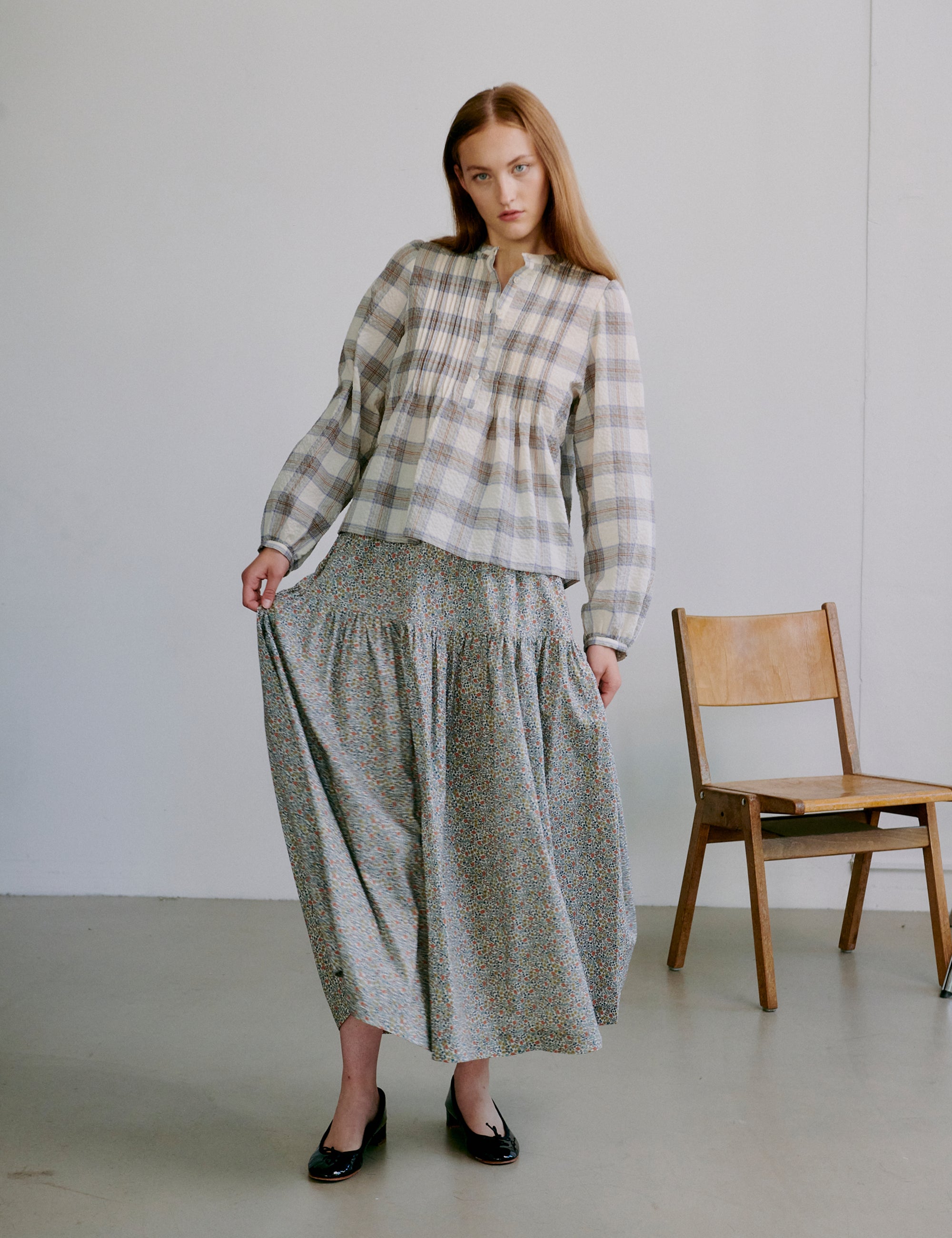 Woman wearing a check patterned white and grey shirt and long floral skirt standing in a minimalistic room with a wooden chair.