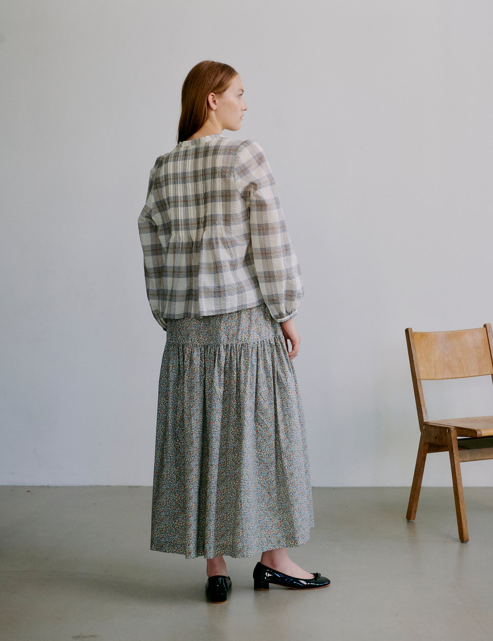 Woman wearing a check patterned white and grey shirt and long floral skirt standing in a minimalistic room with a wooden chair.