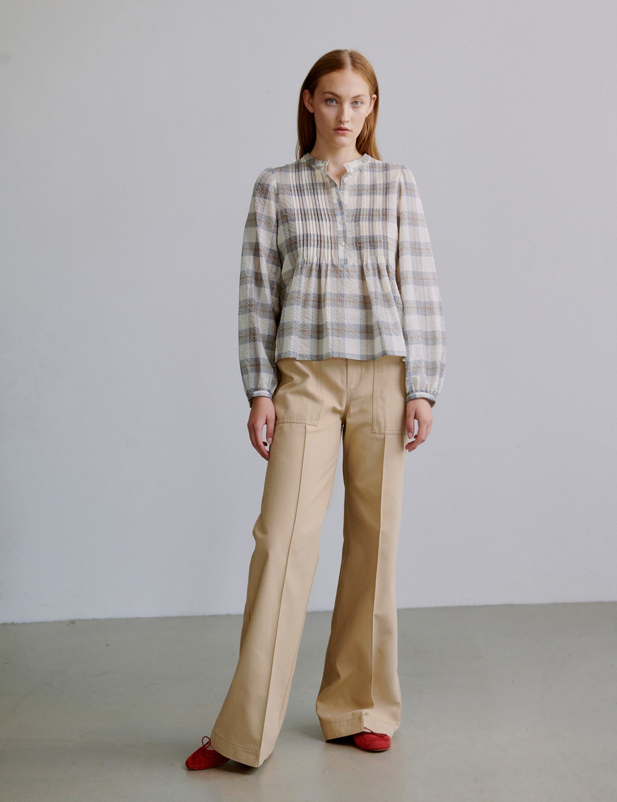 Woman wearing a grey checked shirt and beige wide pants against a plain background