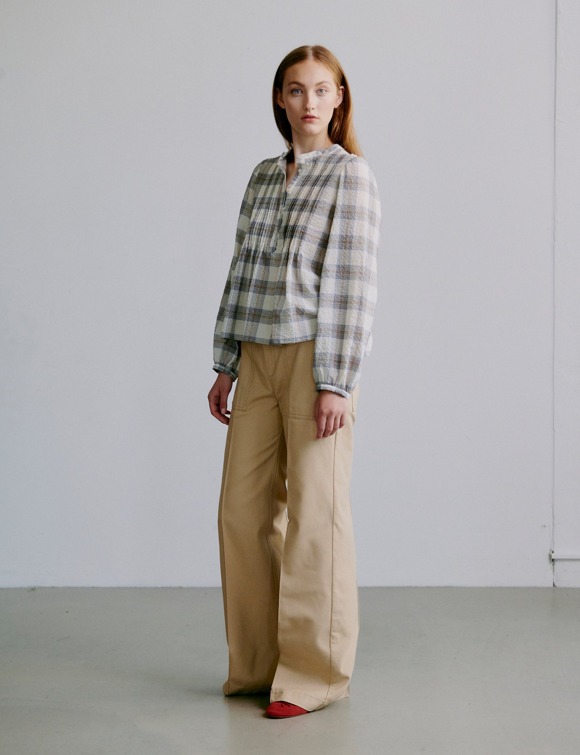 Woman wearing a grey checked shirt and beige wide pants against a plain background