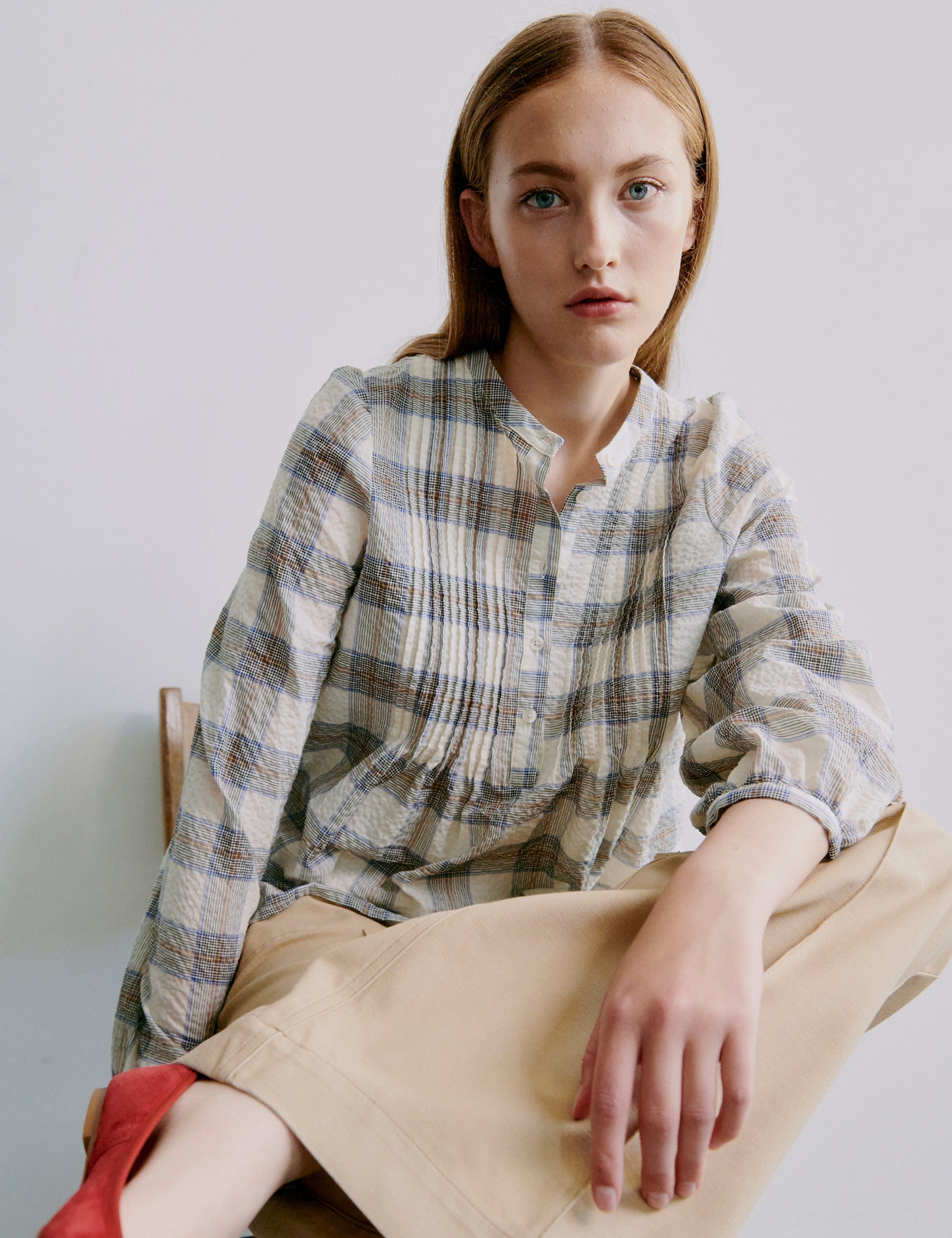 Woman wearing a checked seersucker shirt and beige pants sitting on a chair against a plain background