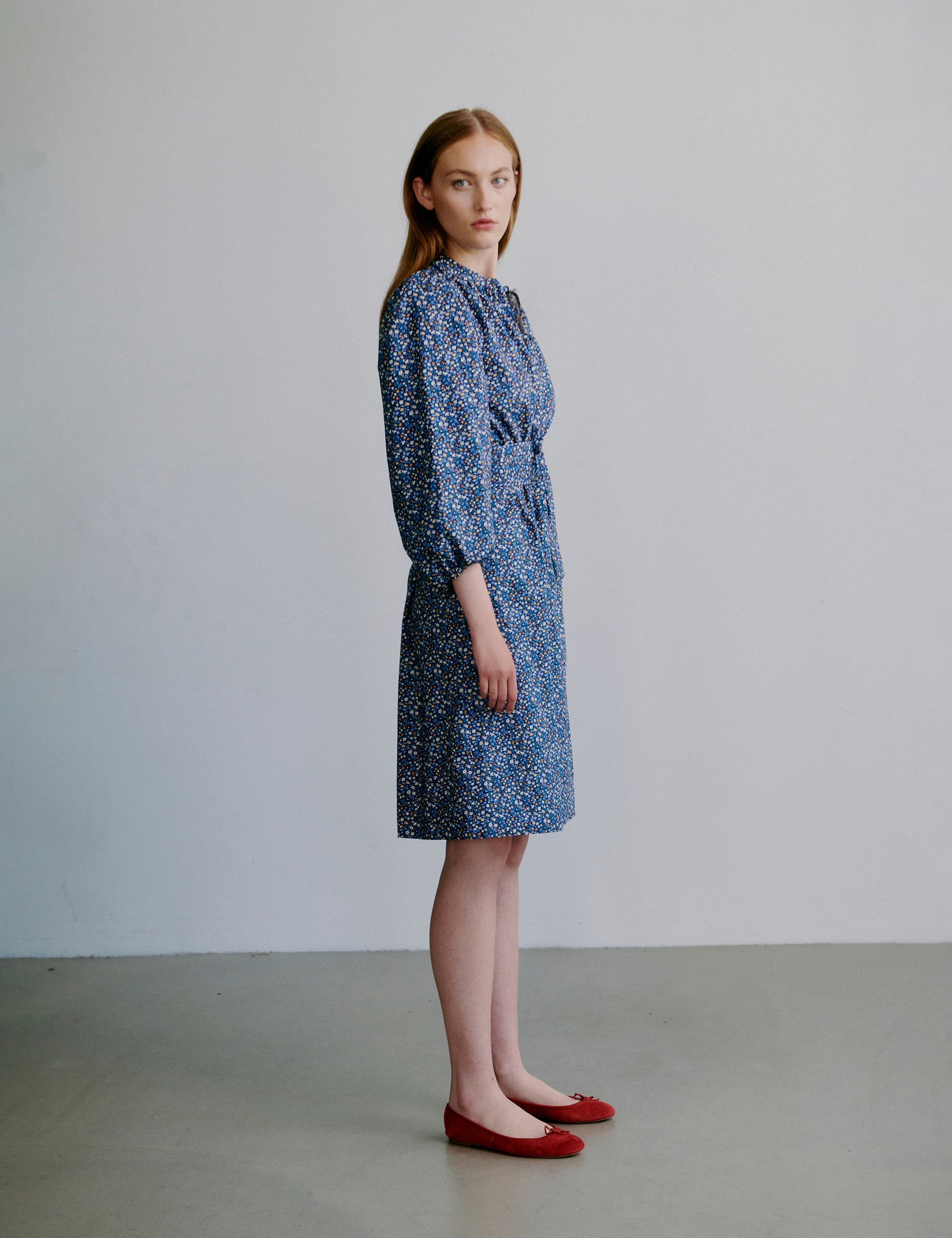 Woman wearing a short blue floral dress with puff sleeves, matching belt and red shoes on a plain background