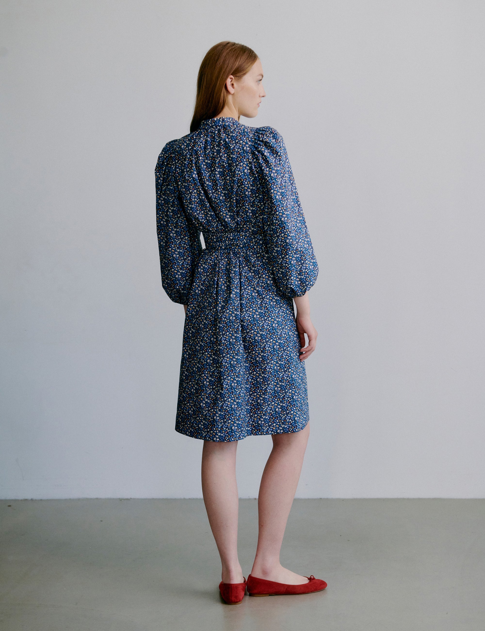 Woman wearing a short blue floral dress with puff sleeves, matching belt and red shoes on a plain background