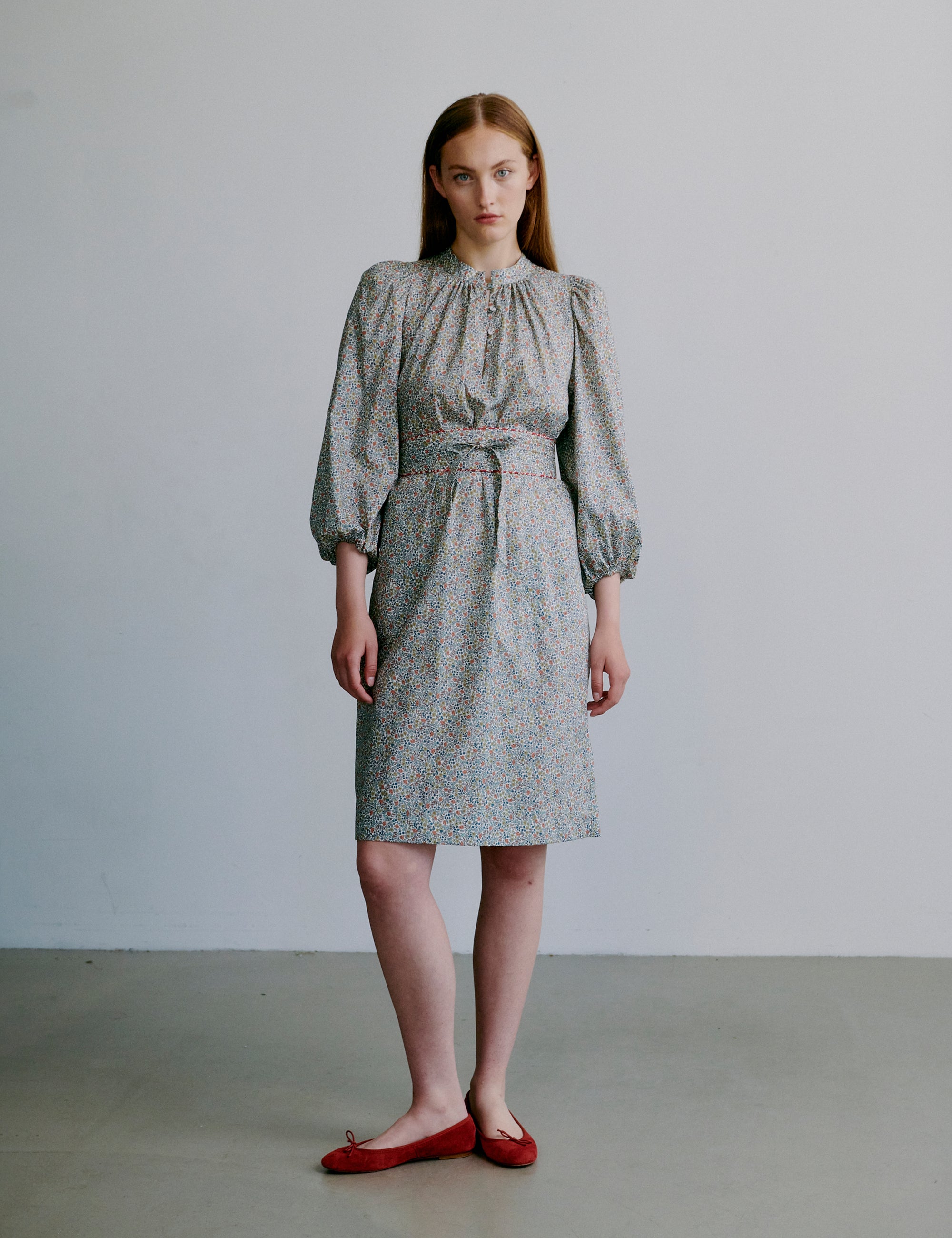 Woman wearing a short floral dress with puff sleeves, matching belt and red shoes on a plain background
