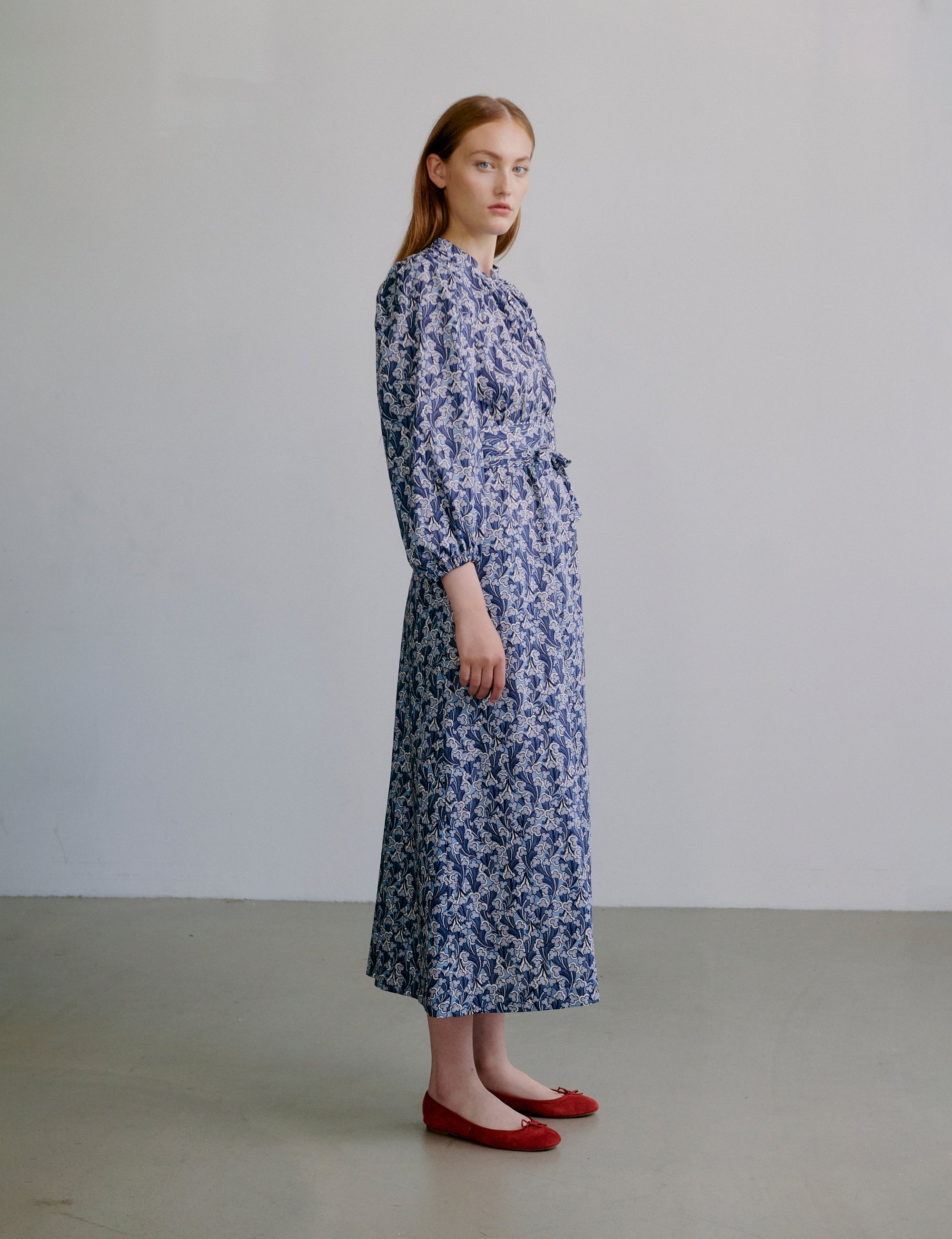 Woman wearing a blue floral dress with long sleeves, matching belt and red shoes against a plain background