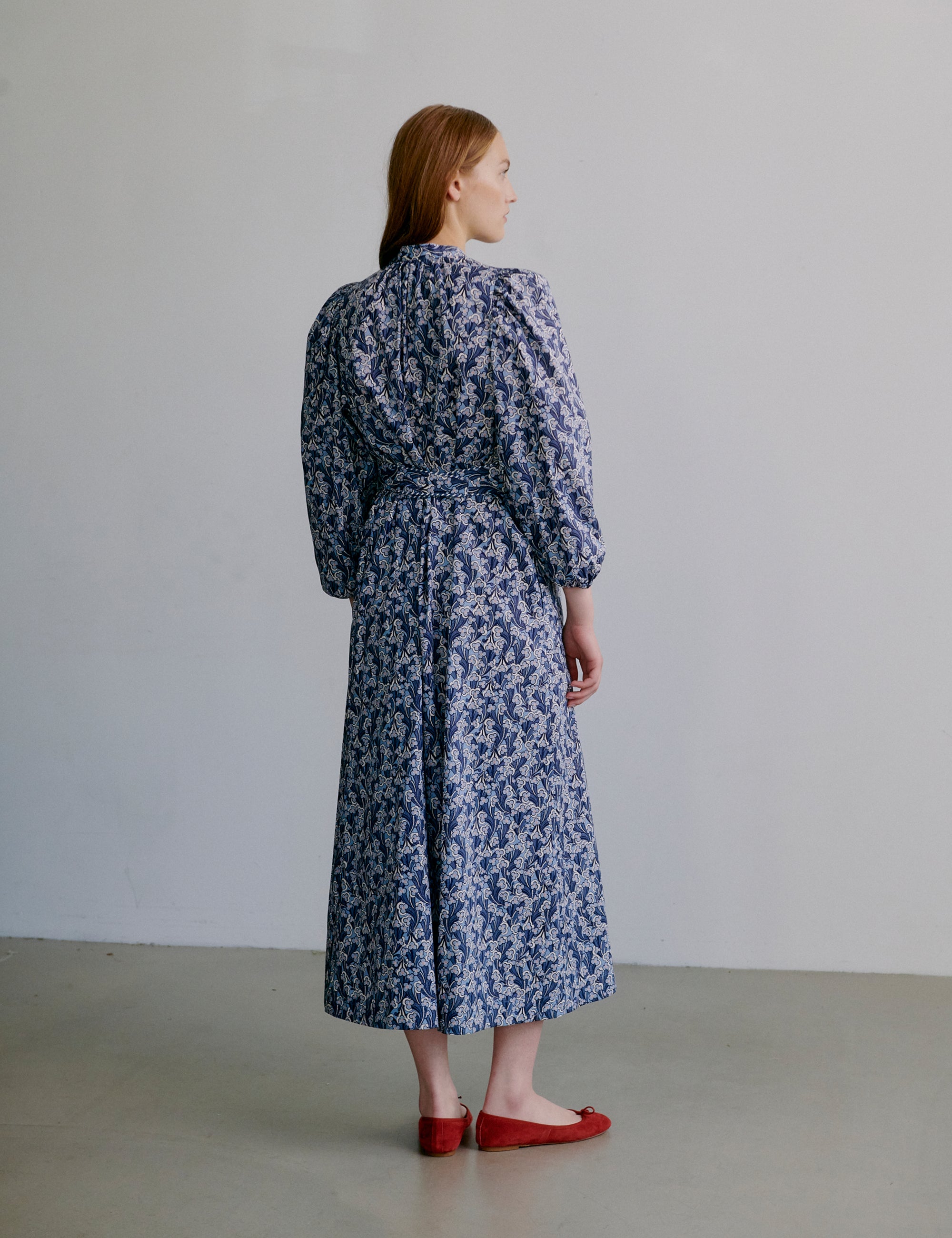 Woman wearing a blue floral dress with long sleeves, matching belt and red shoes against a plain background