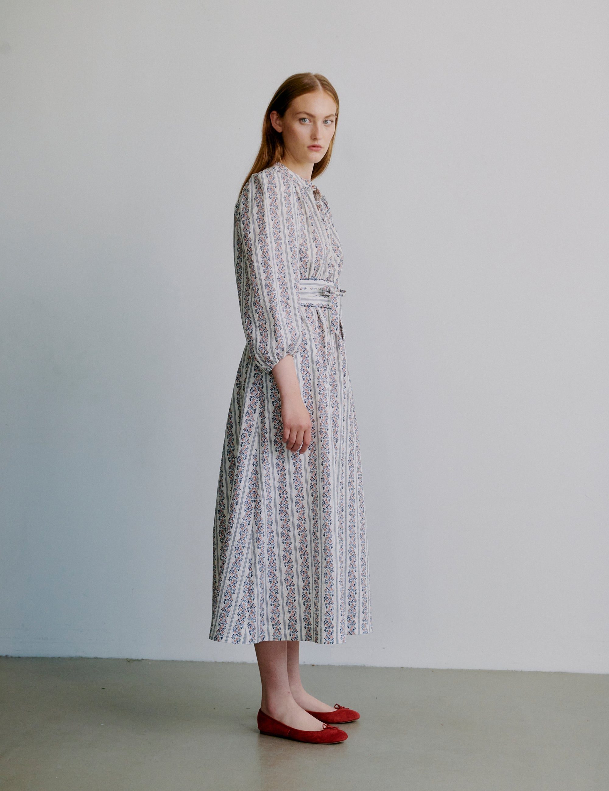 Woman wearing a floral and stripe patterned dress with long puff sleeves and matching belt against a plain background