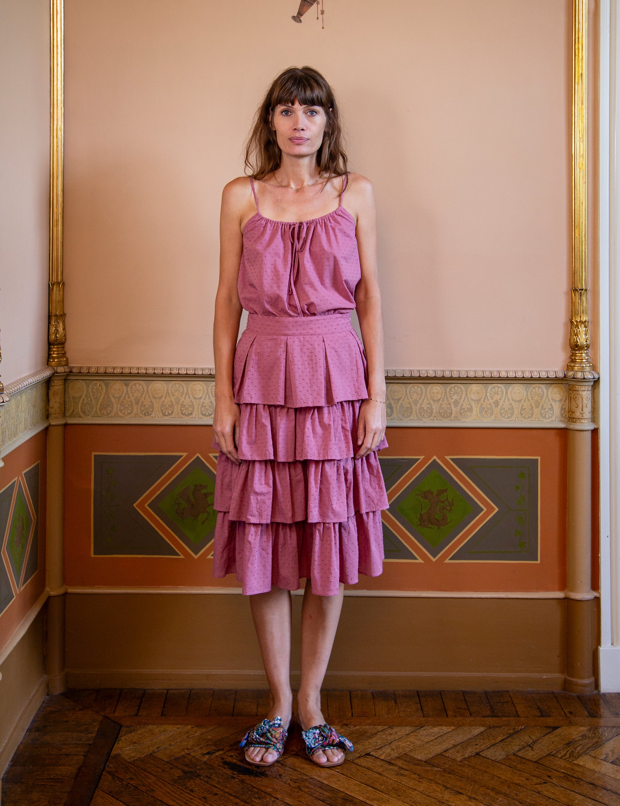 Woman wearing a pink tiered skirt and matching strap top standing in an ornate room with decorative walls.