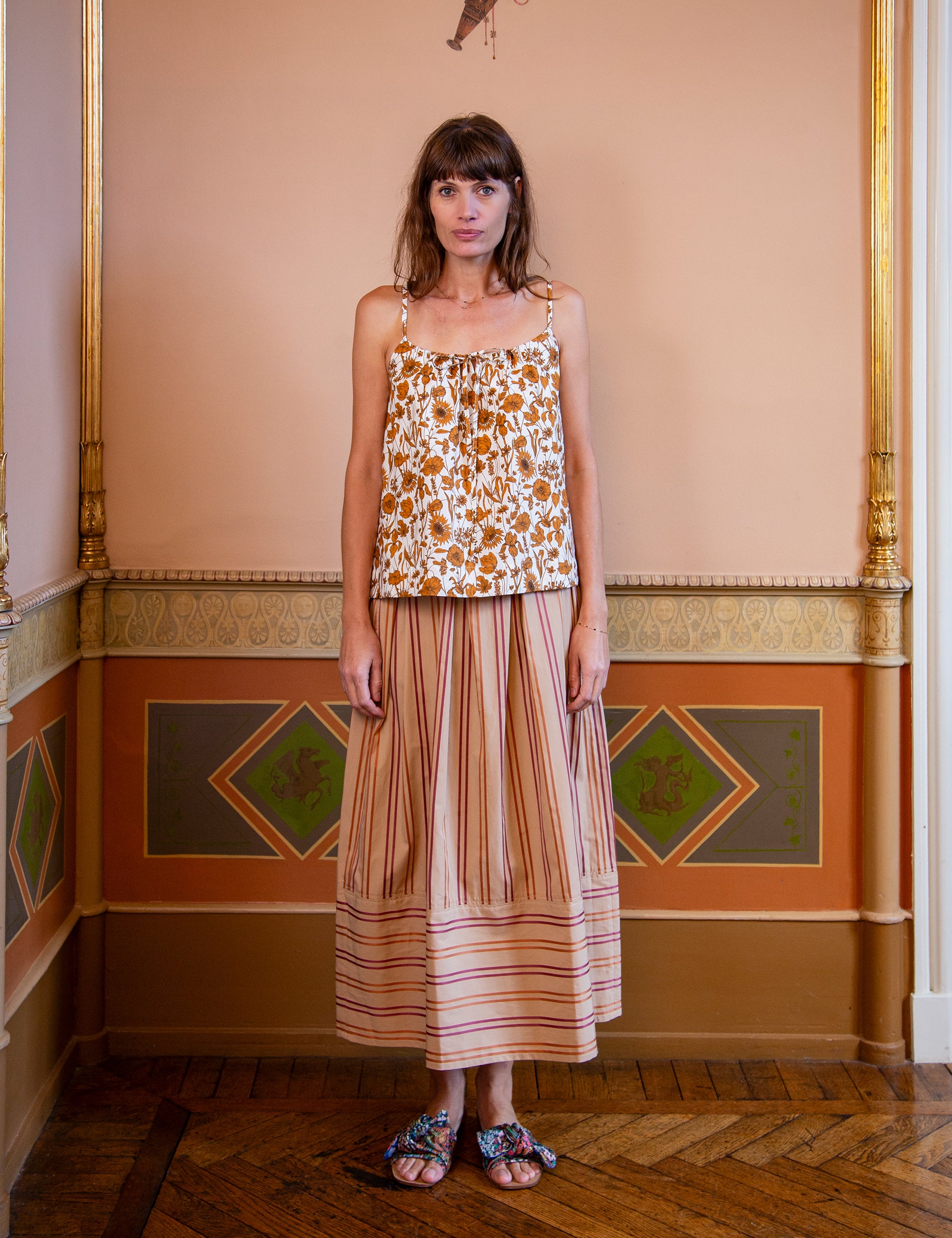 Woman wearing a floral patterned strap top and striped skirt in an ornate room.