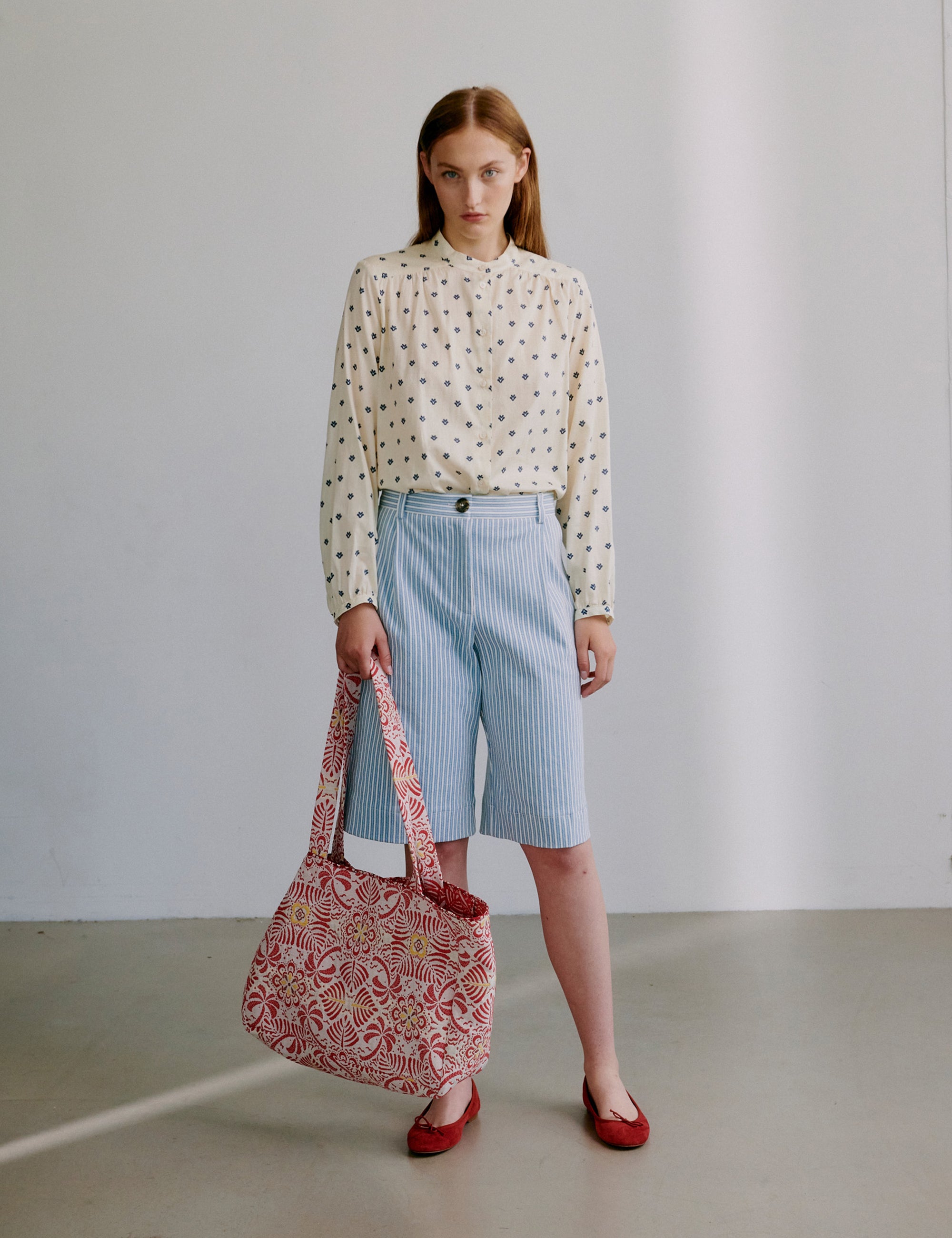 Woman holding a red patterned bag, wearing a dotted blouse and blue striped shorts, against a plain background