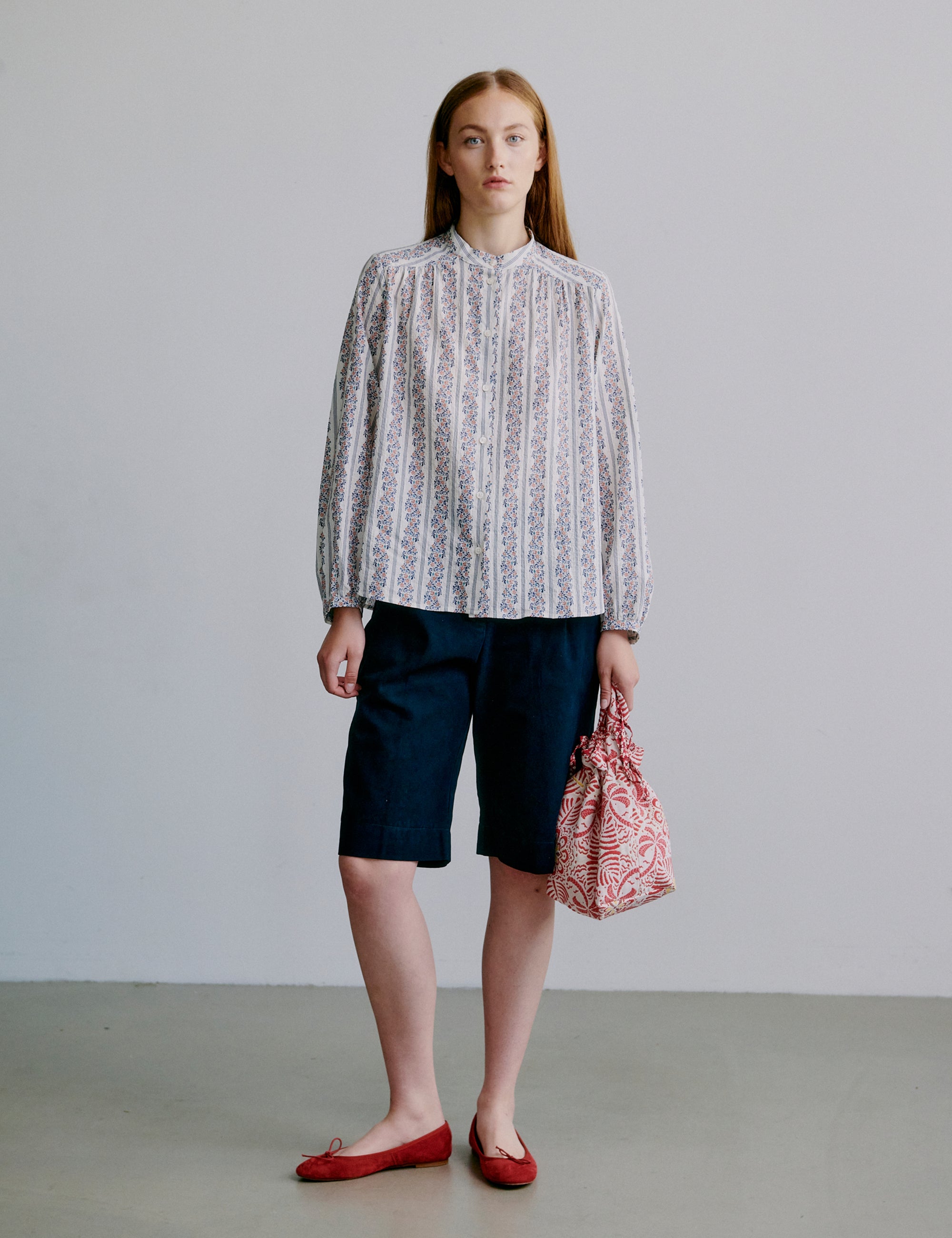 Woman wearing a long sleeve floral patterned blouse and navy shorts, holding a red fabric bag against a plain background
