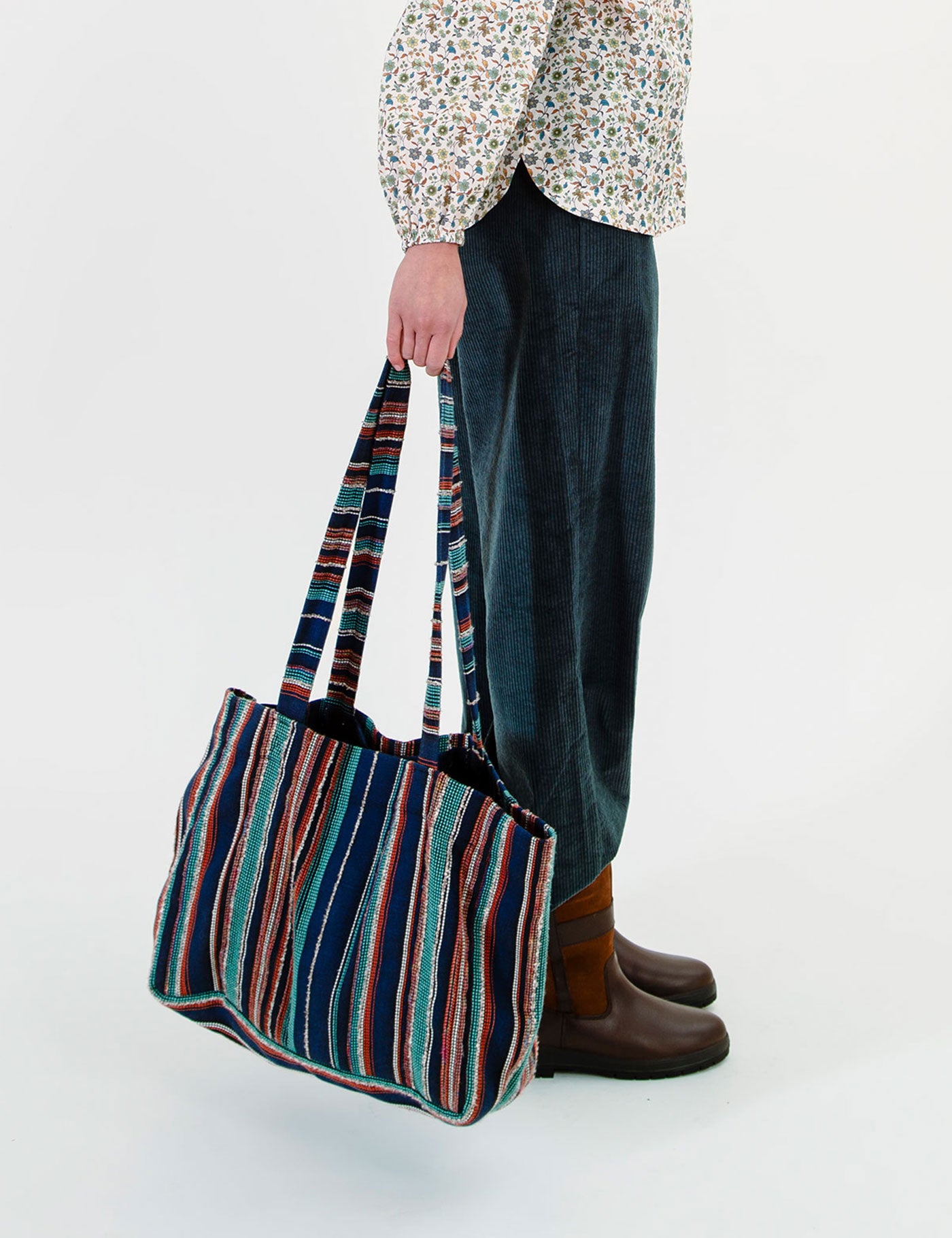 A person wearing a floral blouse, dark blue corduroy pants, and brown boots holds a large striped tote bag with navy, teal, red, and beige vertical stripes against a plain white studio background.