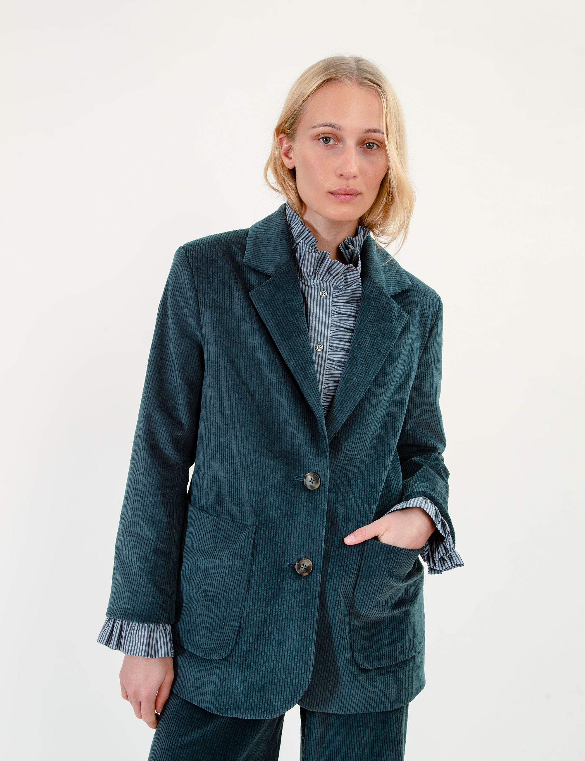 A woman wearing a dark teal corduroy blazer over a blue and white striped shirt, paired with matching corduroy trousers, against a plain white studio background.