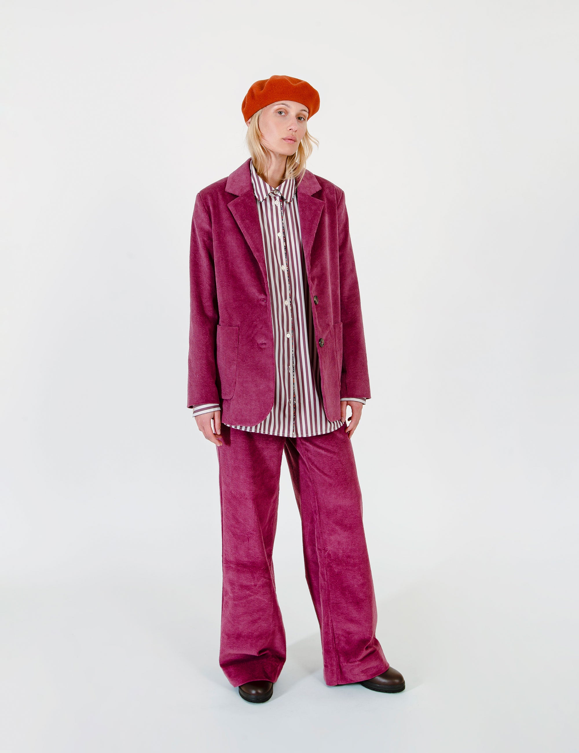 A woman wearing a matching magenta corduroy blazer and trousers set, a vertically striped button-up shirt, and an orange beret.