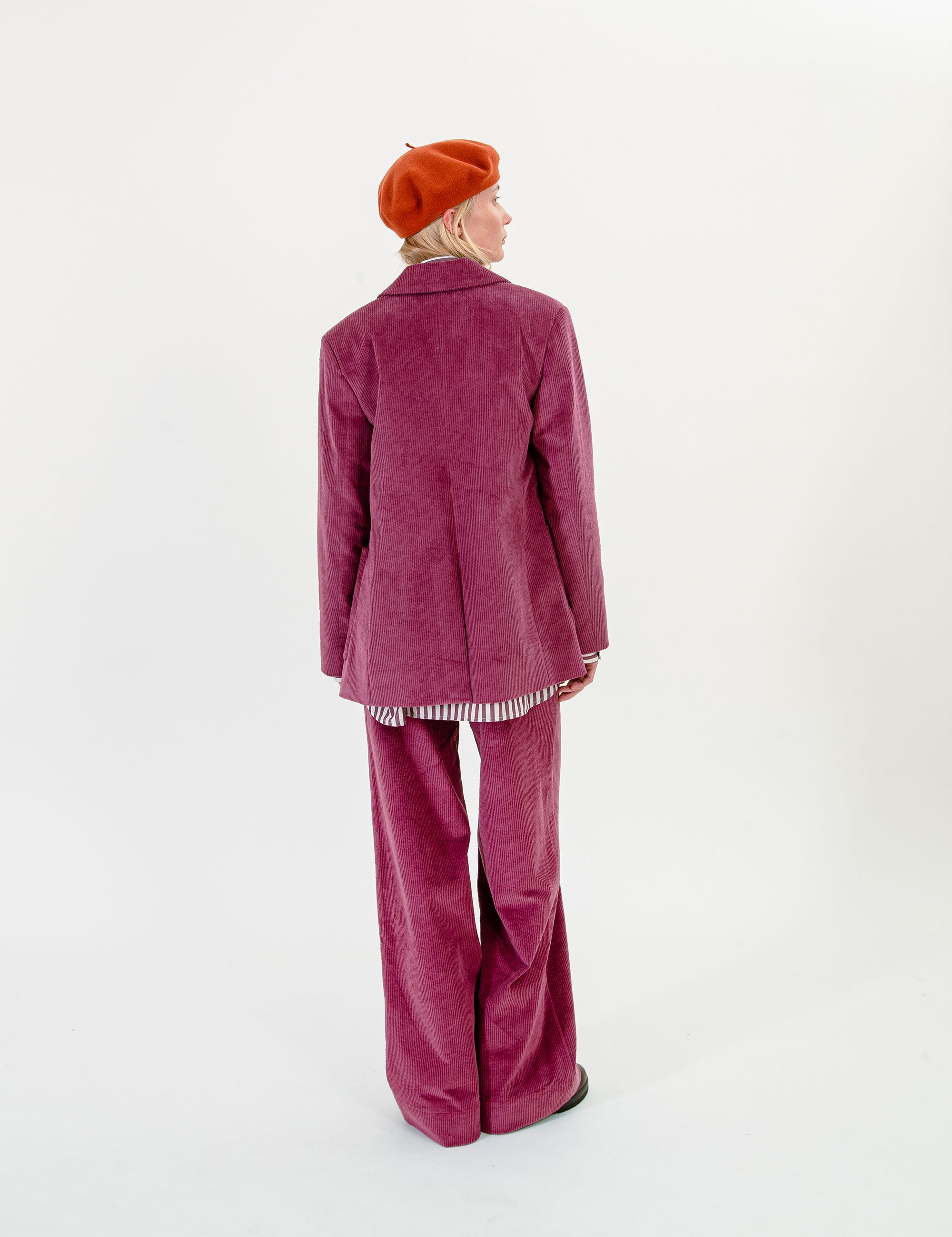 A model shown from the back wearing a matching magenta corduroy blazer and wide-leg trousers, paired with a bright orange beret, against a plain white studio background.