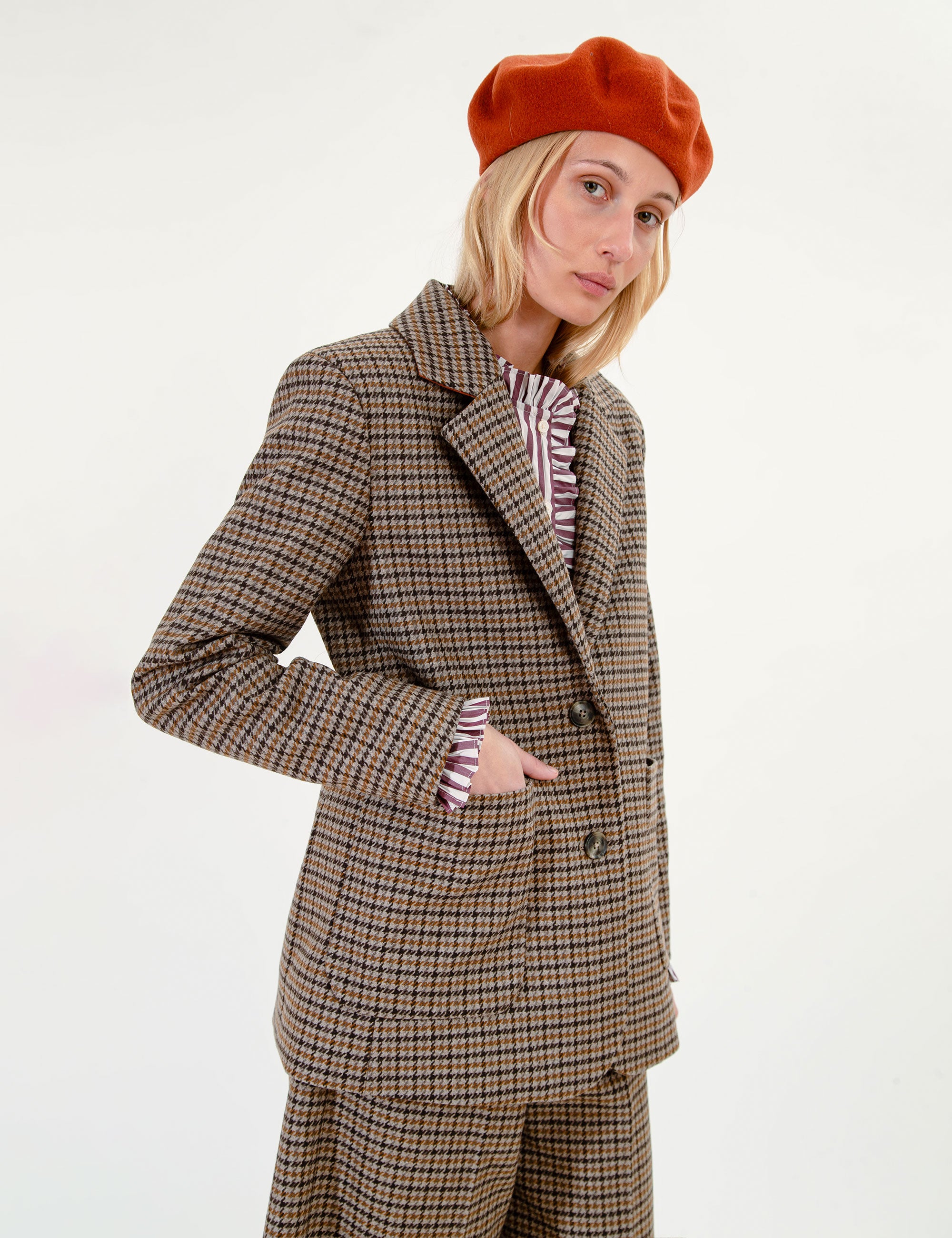A woman stands against a plain white studio background wearing a brown and beige plaid blazer and matching shorts, a striped shirt, a rust-orange beret.
