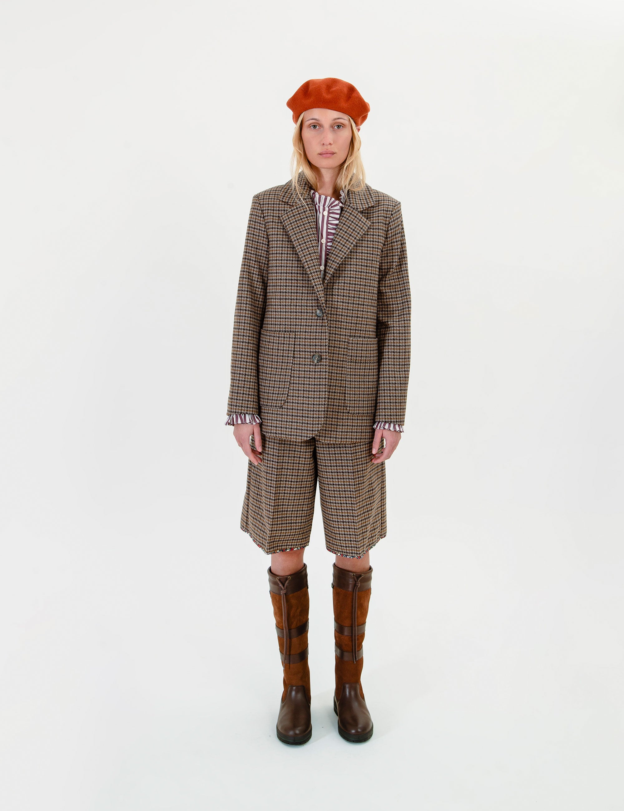 A woman stands against a plain white studio background wearing a brown and beige plaid blazer and matching shorts, a striped shirt, a rust-orange beret, and brown plaid knee-high socks.