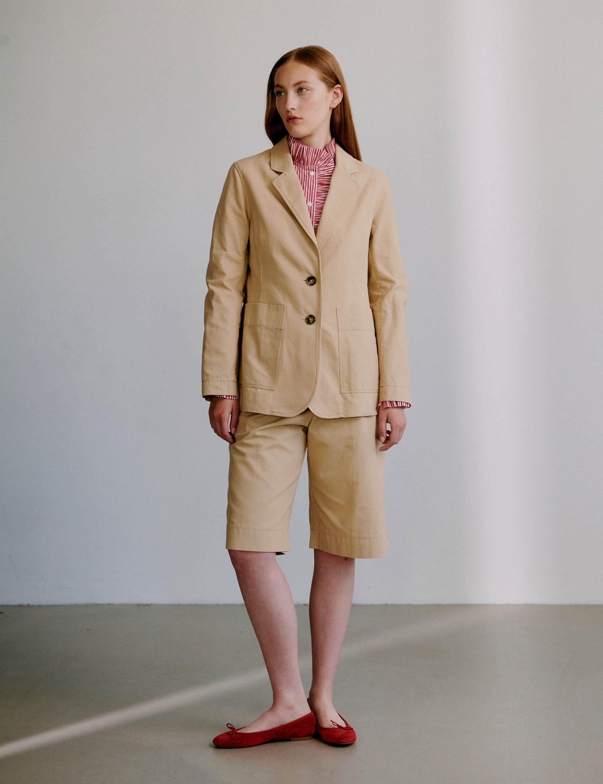 Woman wearing a beige jacket with a red patterned shirt underneath and matching shorts, standing against a plain background.