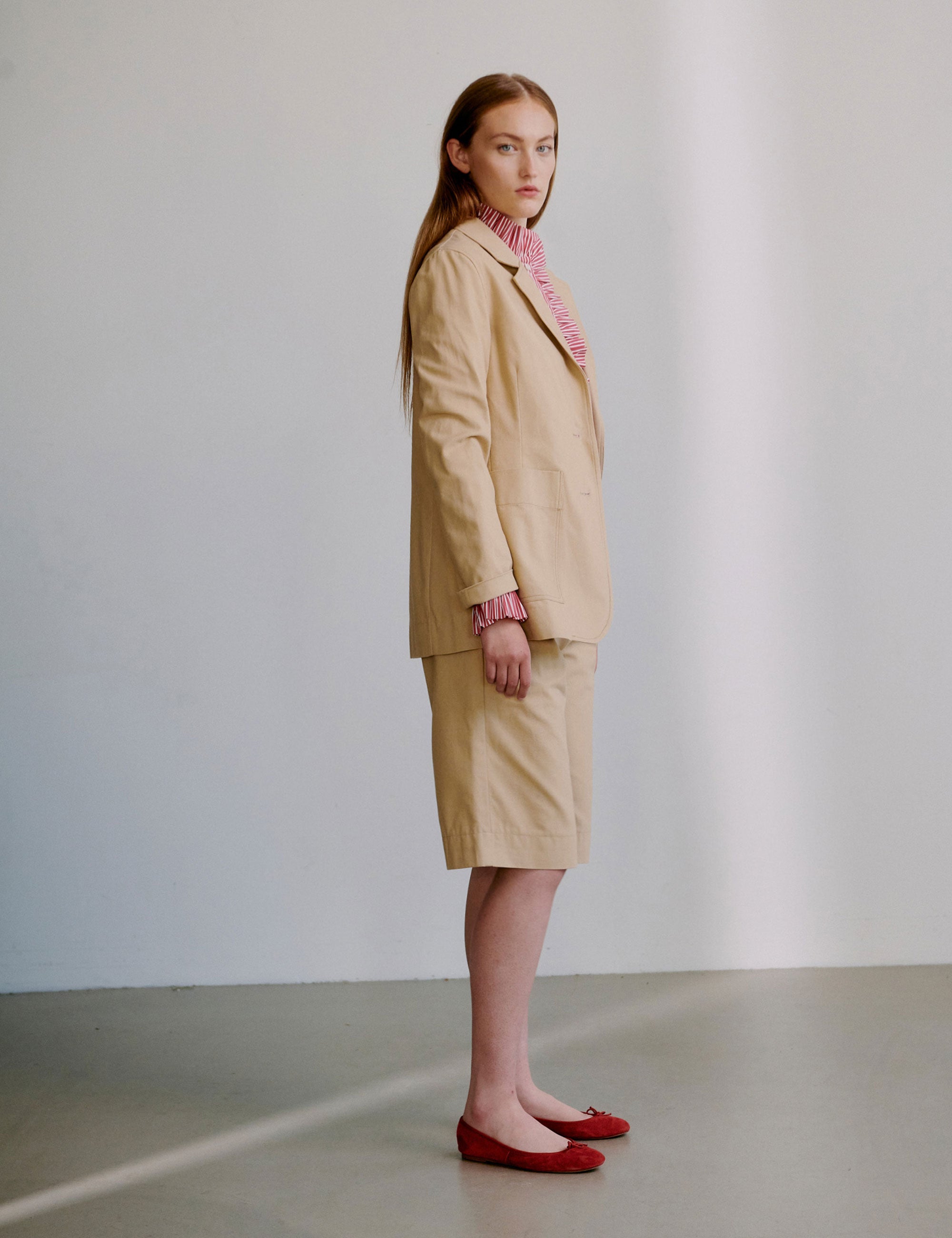 Woman wearing a beige jacket with a red patterned shirt underneath and matching shorts, standing against a plain background.