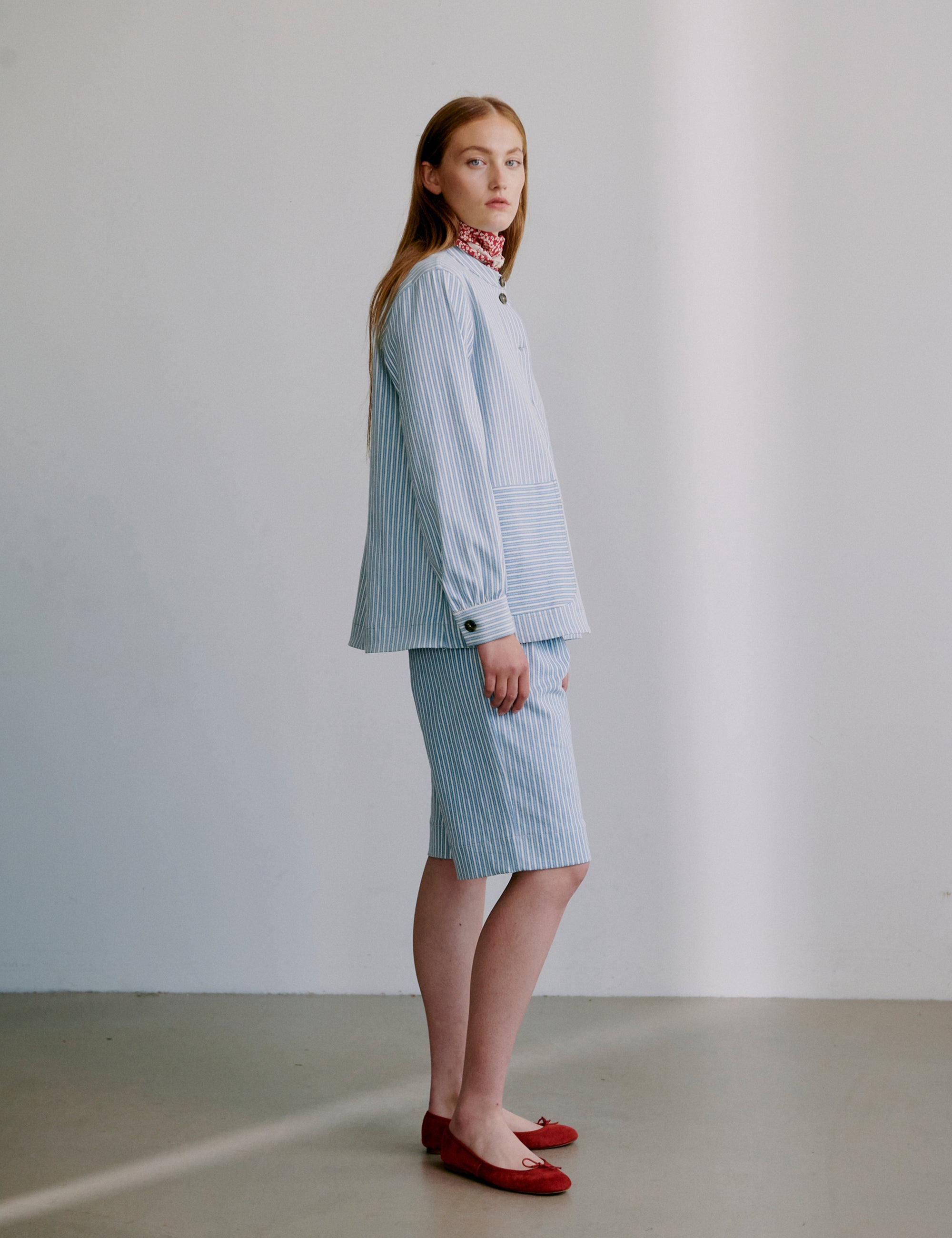 Woman wearing a light blue striped jacket with matching shorts and red shoes on a plain background