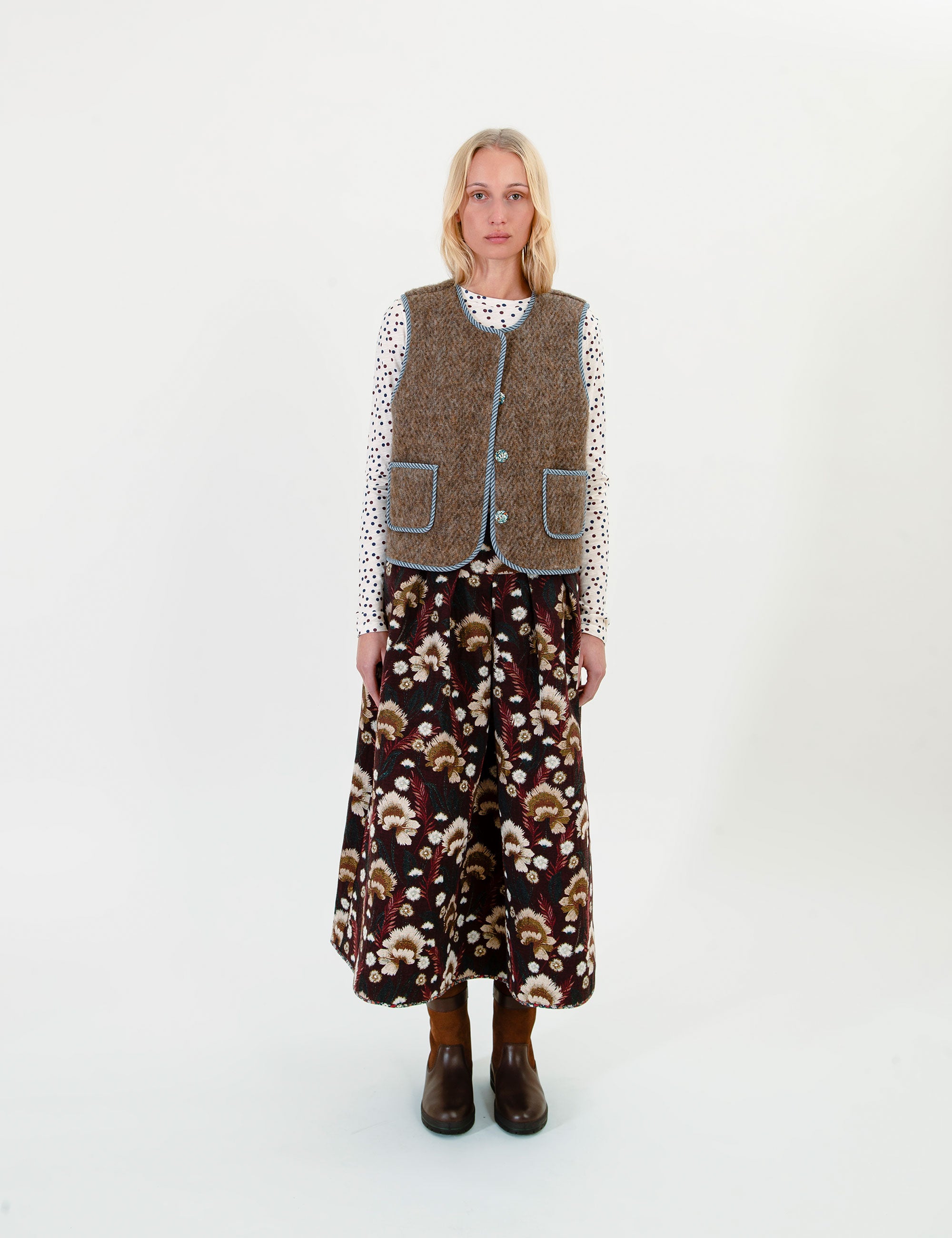 A woman stands against a plain white studio background wearing a brown wool vest over a white patterned long-sleeve top and a dark brown floral midi skirt, paired with dark brown boots.