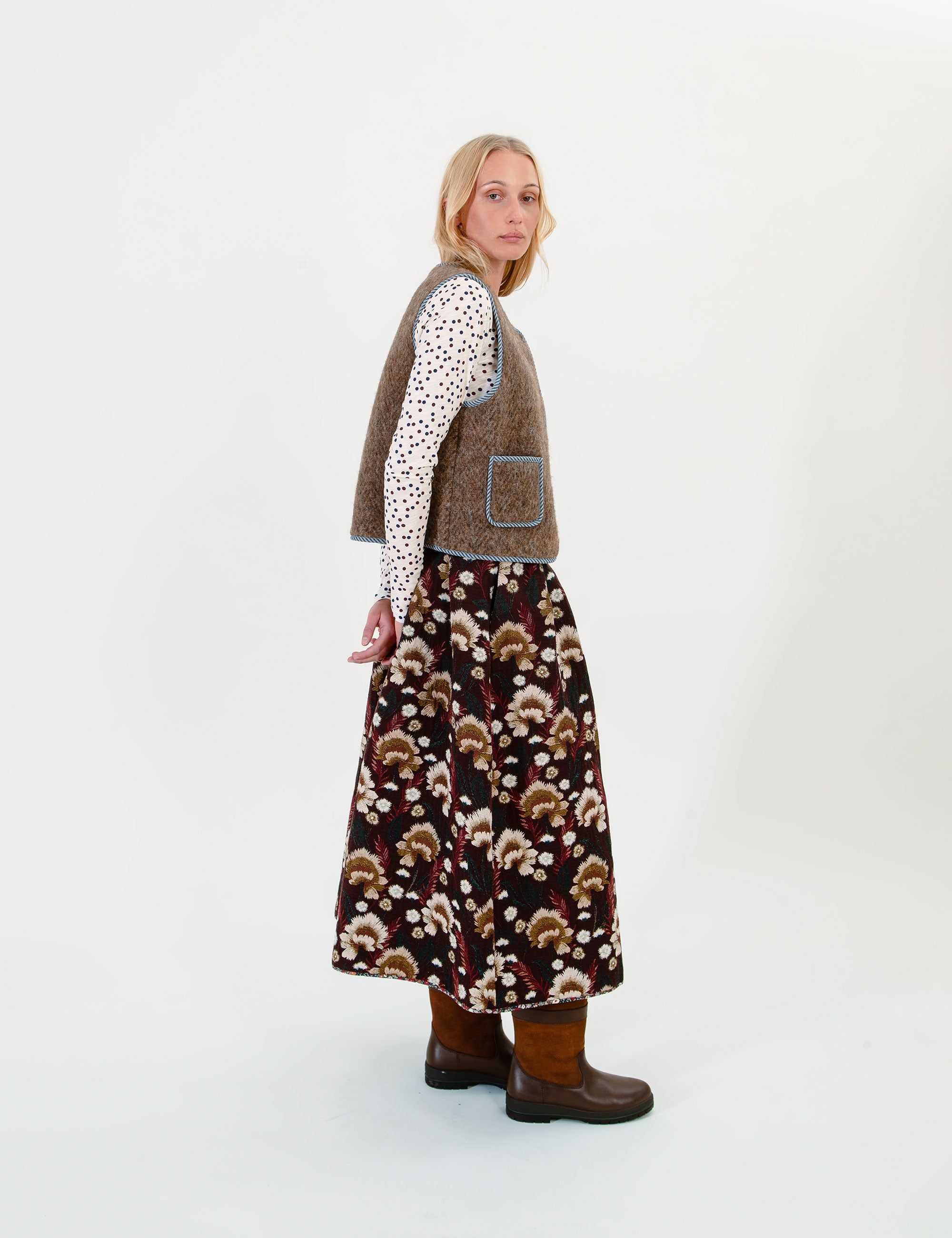 A woman stands in profile wearing a brown vest over a white long-sleeve top with a black polka-dot pattern, a long brown skirt with a brown and cream mushroom print, and brown boots, against a plain white studio background.