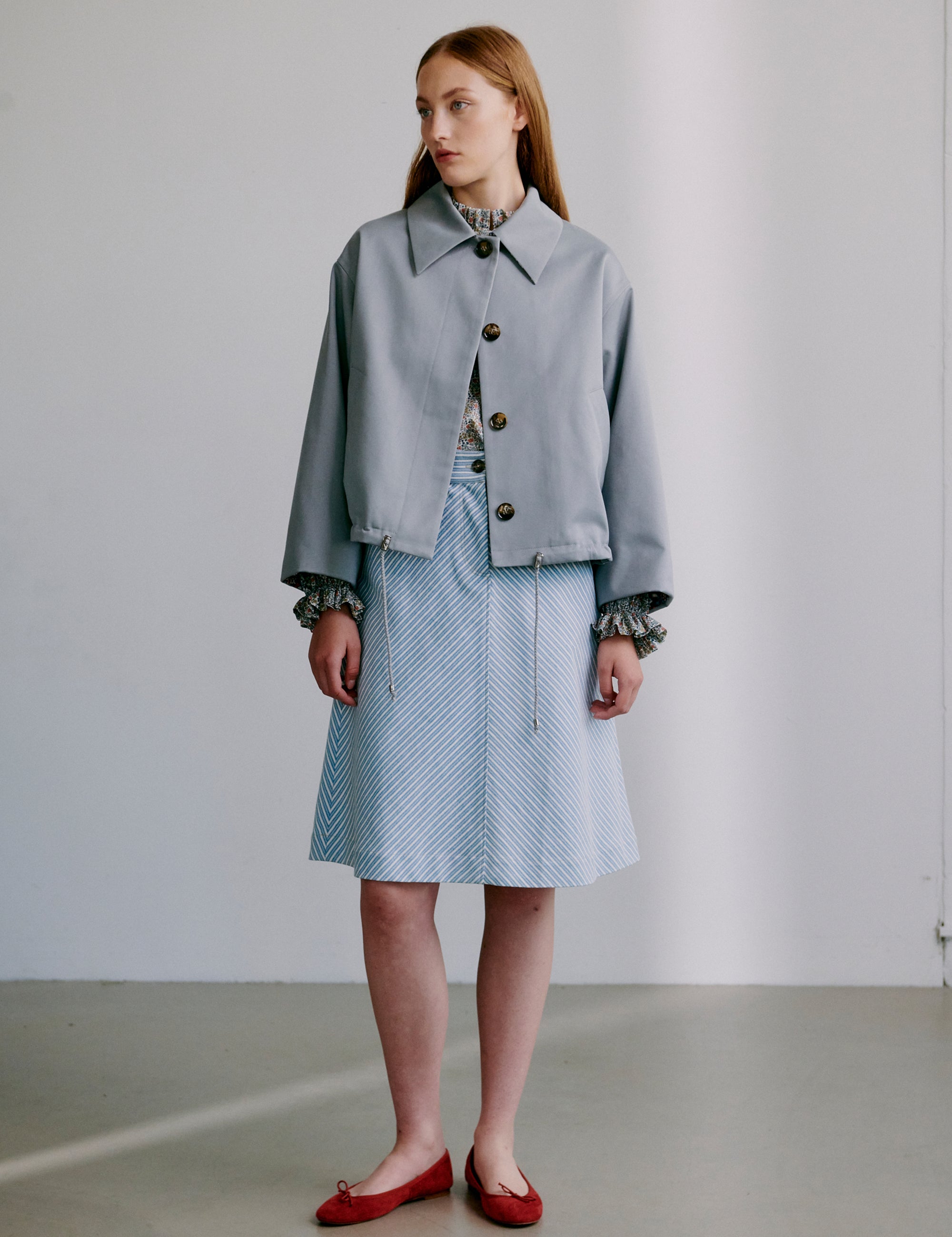 Woman wearing a light grey jacket and blue and white striped skirt with red shoes against a plain background