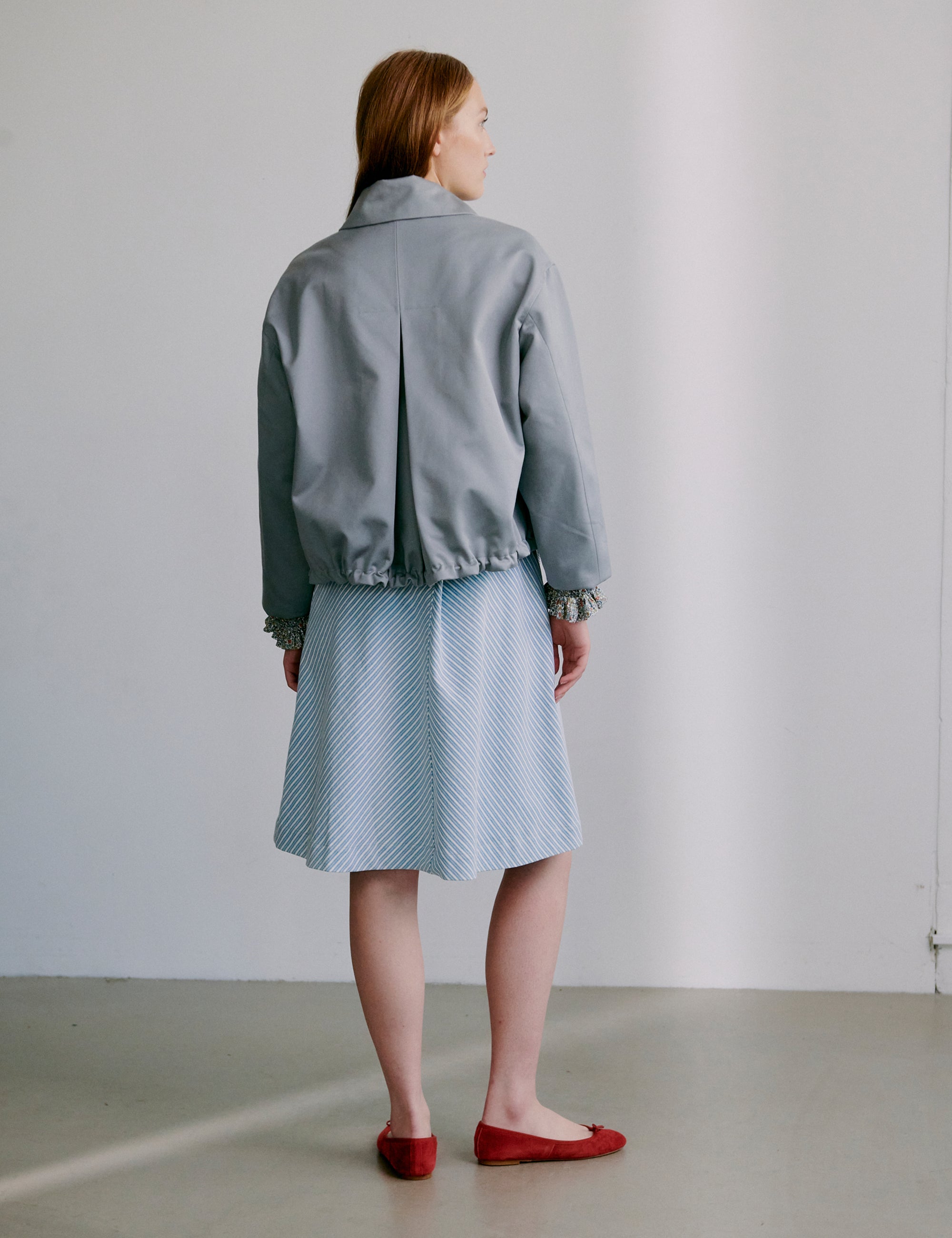 Woman wearing a light grey jacket and blue and white striped skirt with red shoes against a plain background