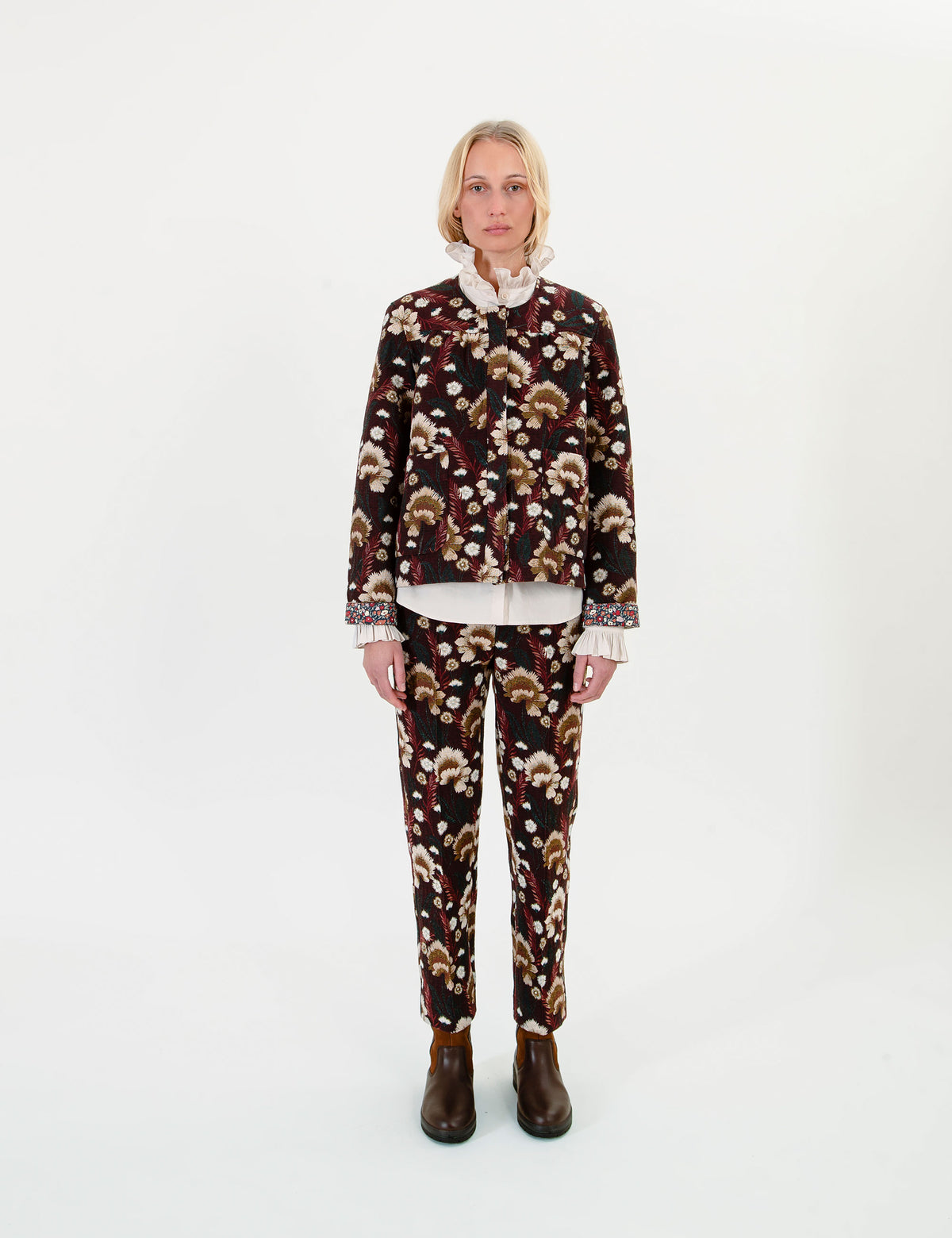 A woman wearing a matching two-piece set consisting of a cropped jacket and trousers with a dark brown and black floral pattern, a white ruffled collar, and white ruffled cuffs, paired with dark brown boots.