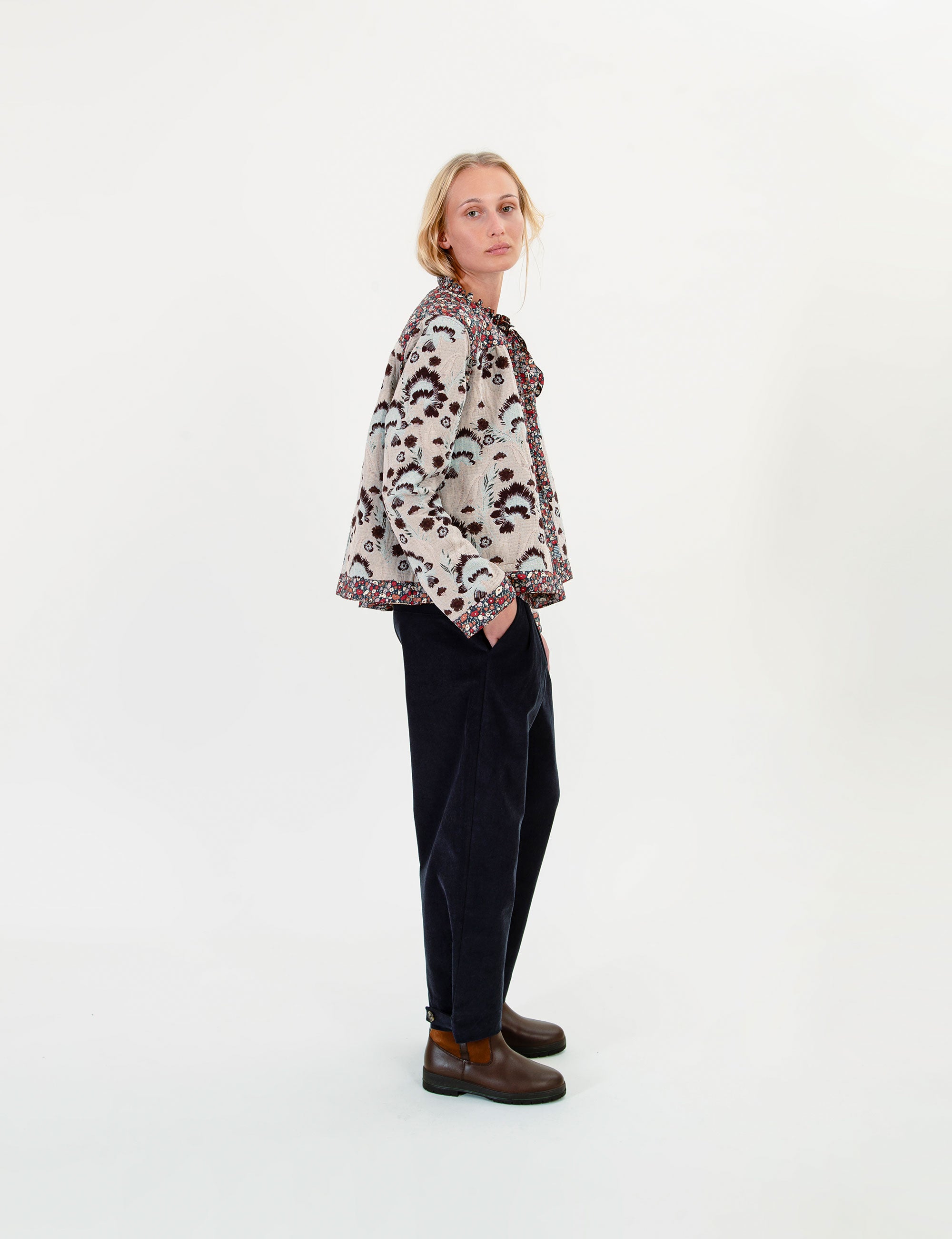 A woman stands in profile wearing a cream patterned jacket with dark trim, dark trousers, and brown boots against a plain white studio background.