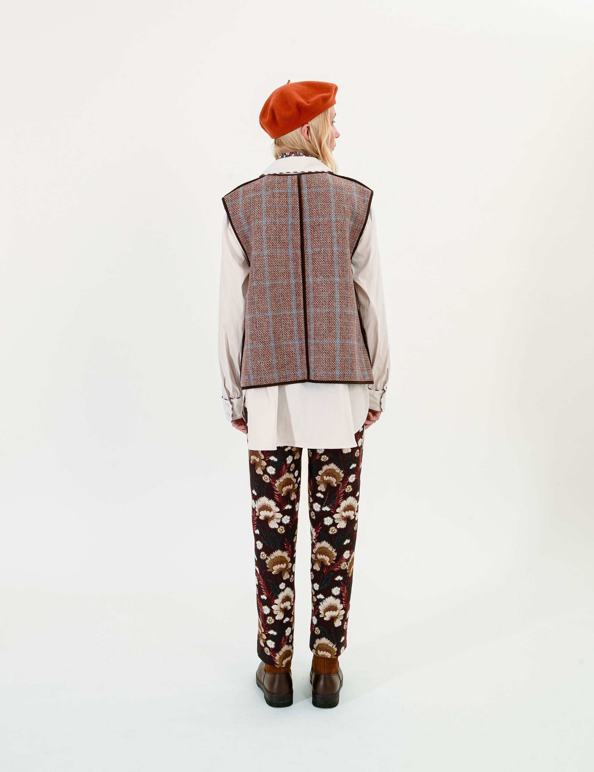 A woman shown from the back wearing a brown and beige plaid vest over a cream shirt, paired with dark brown pants featuring a floral pattern and an orange beret, against a plain white studio background.