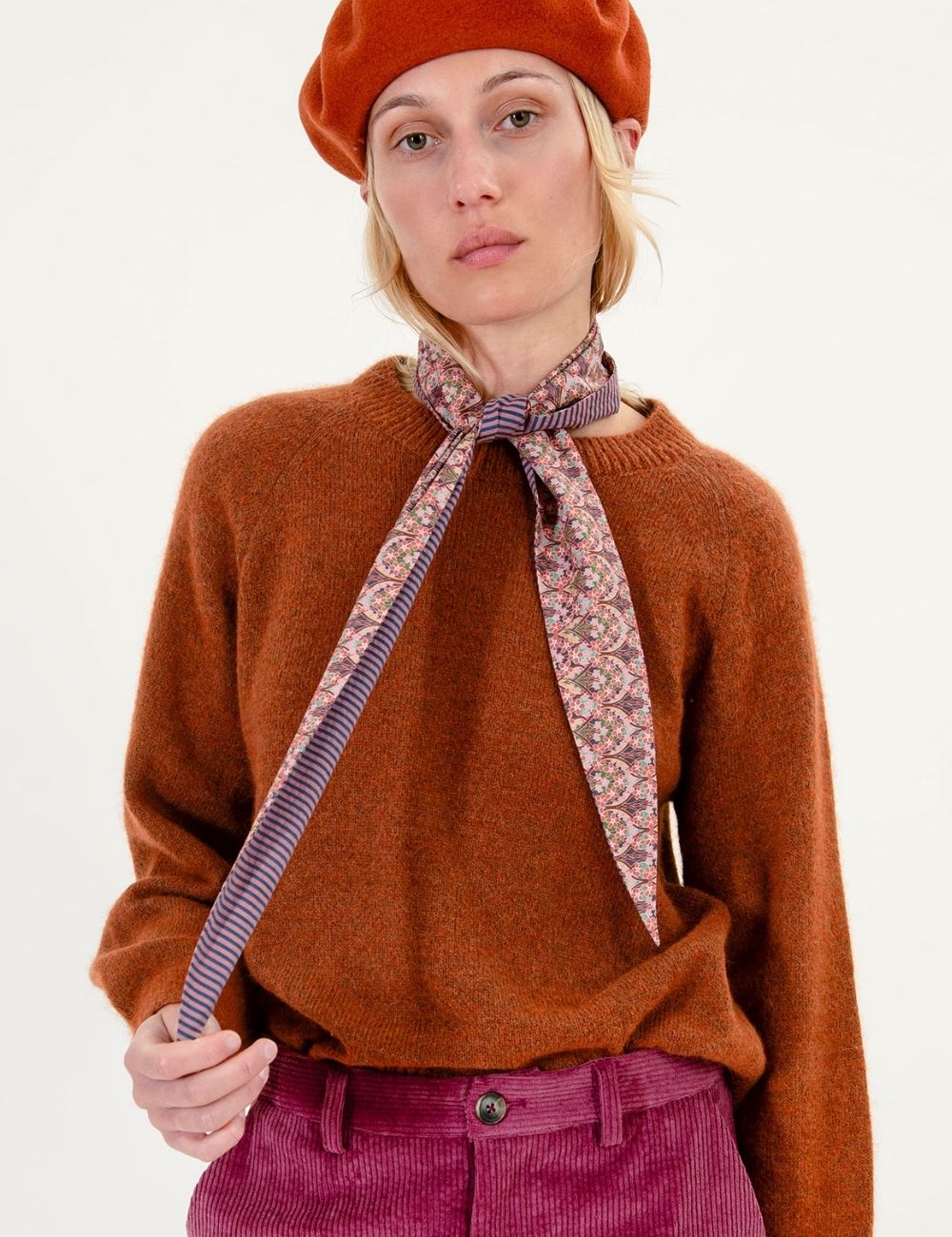 A woman wearing a rust-orange knit sweater, a matching rust-orange beret, and a purple patterned neck scarf tied at the collar, paired with magenta corduroy trousers against a plain white background.