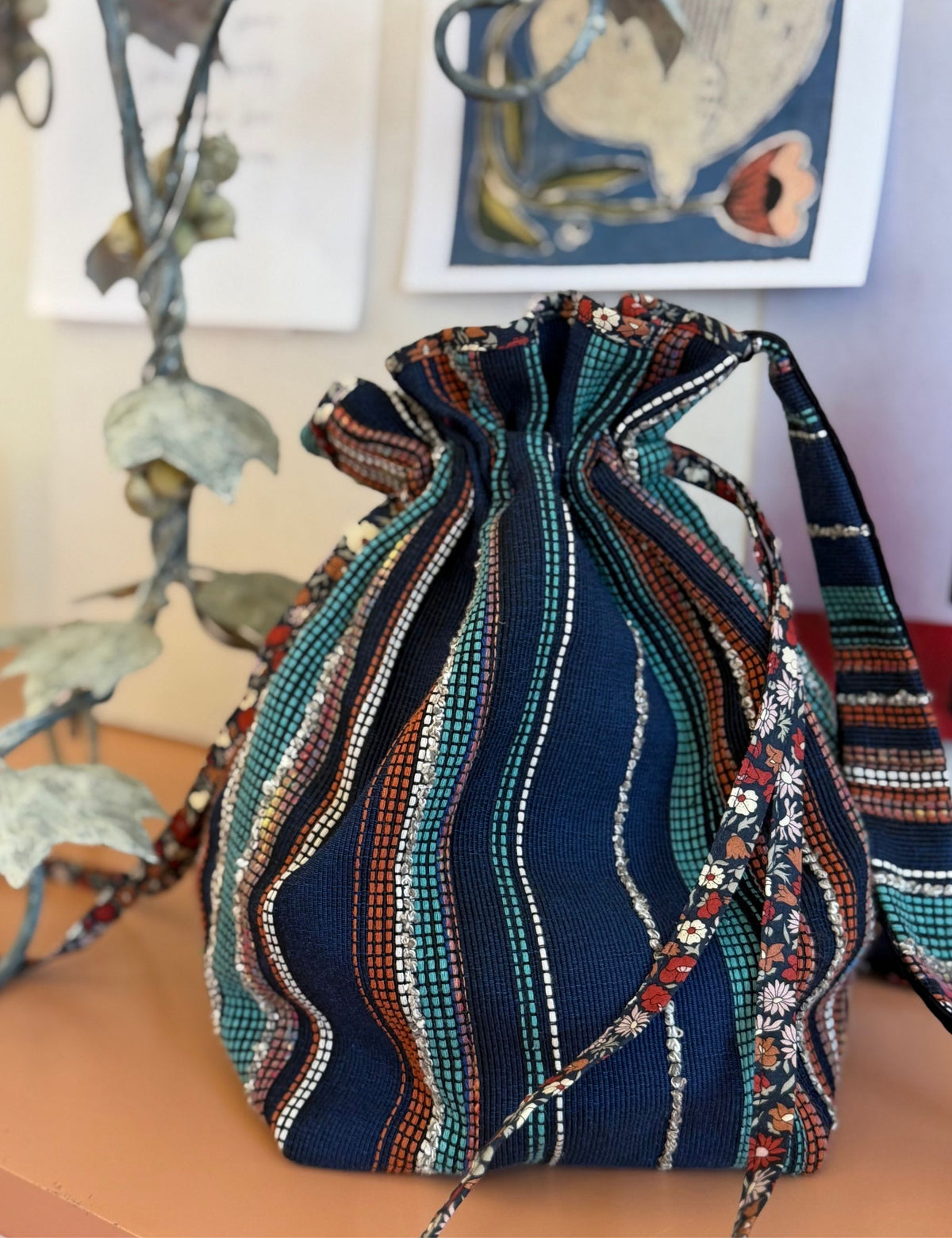 A handmade drawstring pouch made from navy blue woven fabric with vertical stripes of teal, orange, red, and white, displayed on a wooden surface with a decorative metal plant in the background.