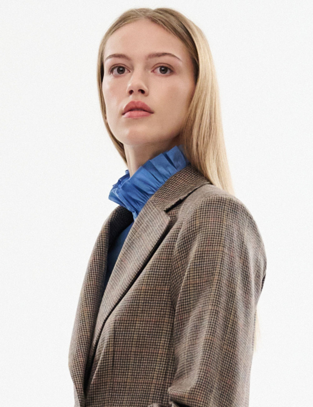 En blond model iført en brun og beige hundetandsmønstret blazer over en lyseblå skjorte med flæser, fotograferet mod en ren hvid studie baggrund.