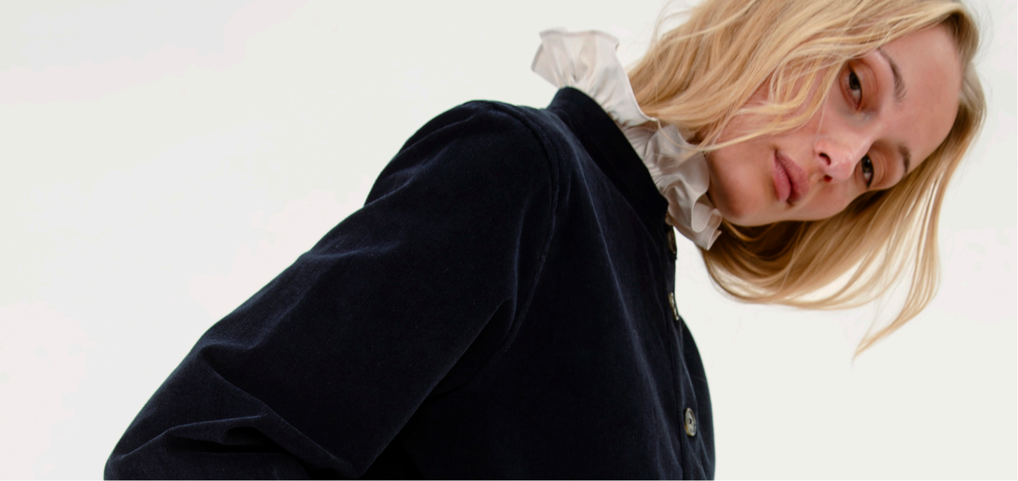 A blonde woman wearing a dark navy velvet jacket over a white ruffled collar blouse, photographed against a clean white background.