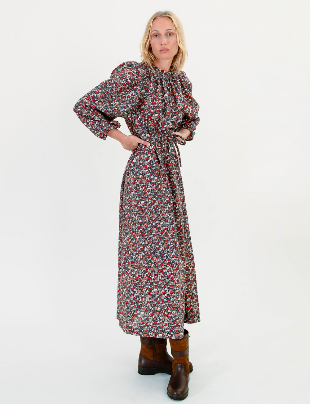 A woman modeling a long-sleeved midi dress with a dense multicolor floral print on a brown base, featuring a high gathered neckline and a self-tie waist belt. She is wearing brown leather boots against a plain white studio background.