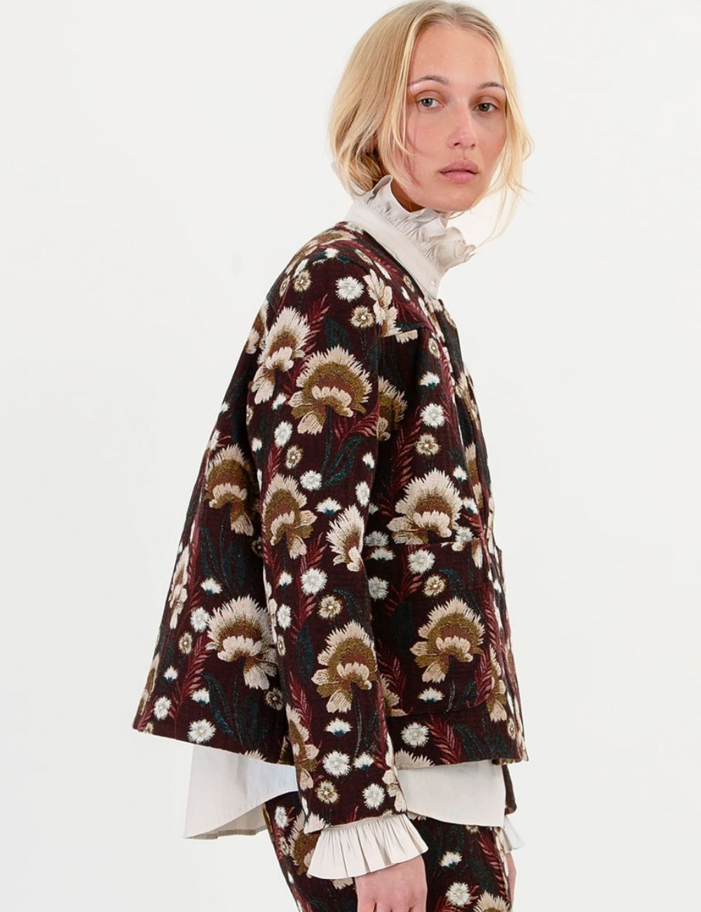 A model wearing a dark brown cropped jacket with a beige and white floral pattern, layered over a white ruffled mock neck top, photographed against a plain white background.