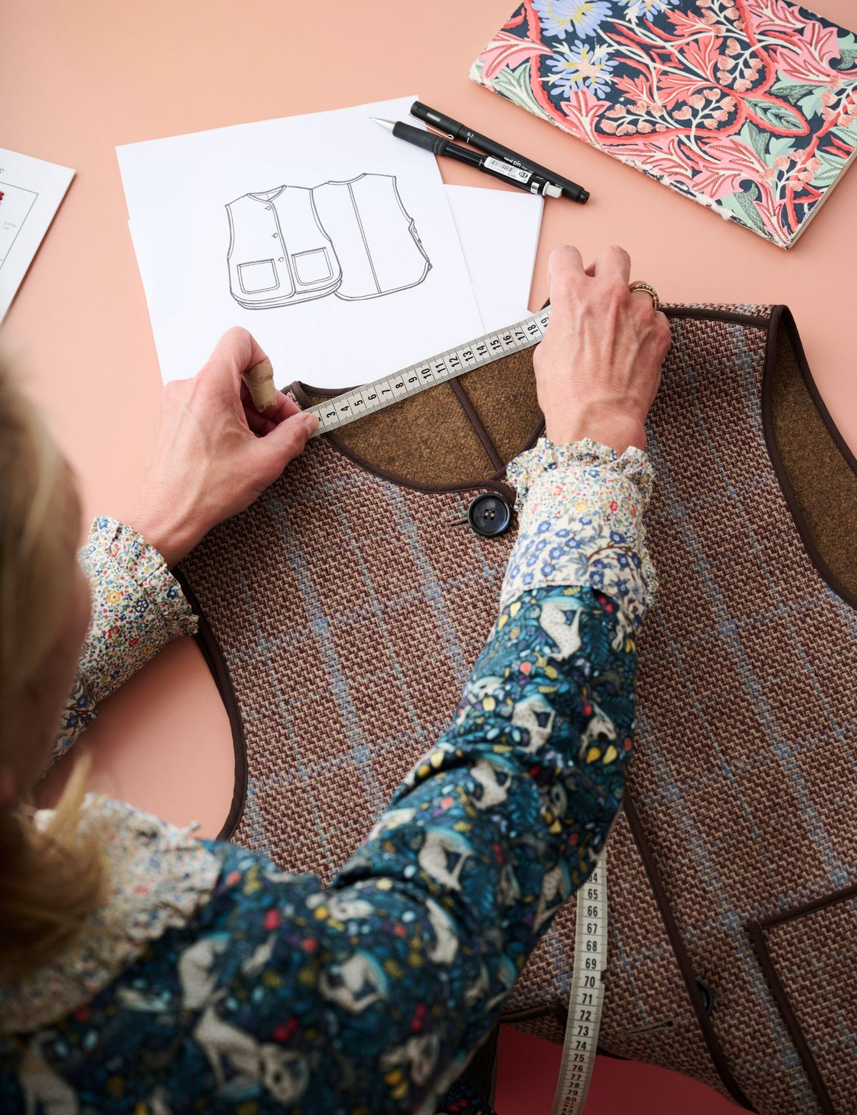 A person measures a brown tweed vest with a white tape measure on a peach-colored desk, with a sewing pattern sheet, pens, and a floral fabric swatch nearby.