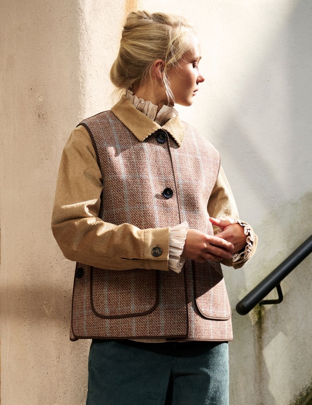 A woman wearing a brown and beige tweed vest over a tan long-sleeve shirt and blue jeans, standing against a textured beige wall.
