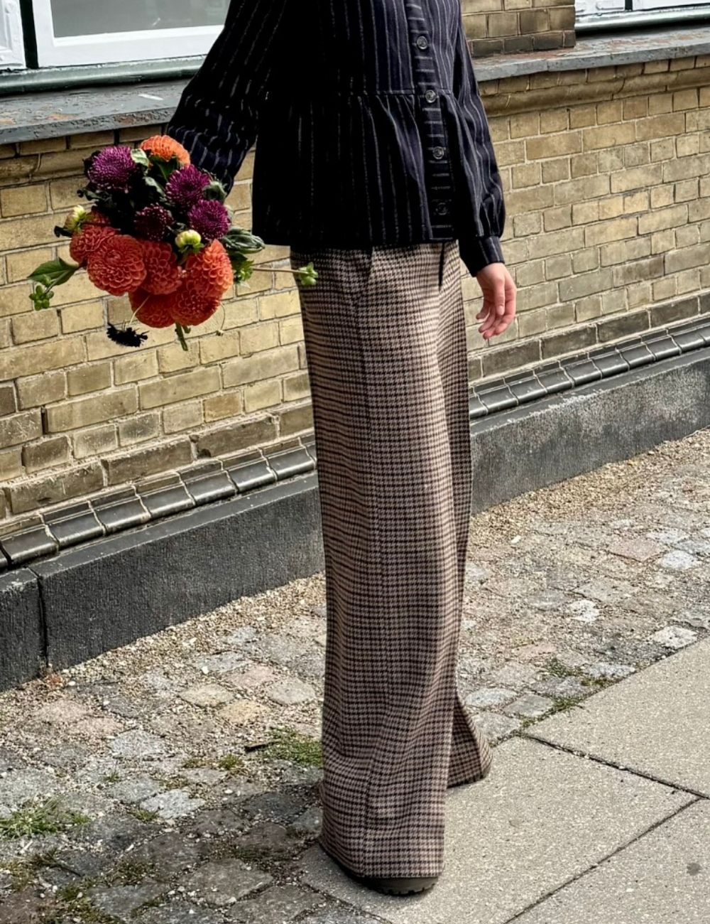 A woman standing outdoors on a sidewalk in front of a brick wall, wearing a dark pinstriped long-sleeve top and a brown and black checkered wide-leg skirt. The person is holding a bouquet of orange, purple, and burgundy flowers.