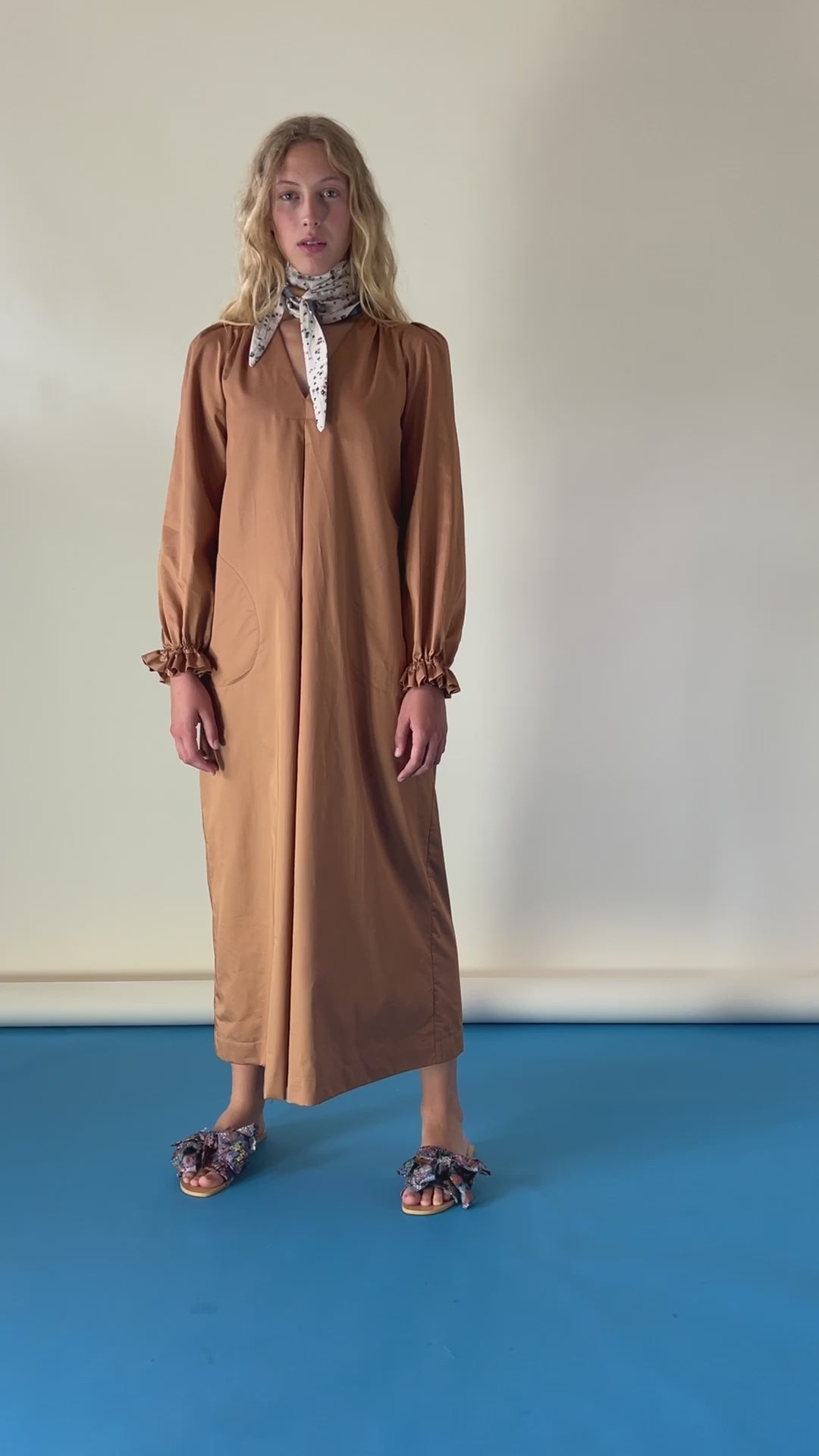 Woman in long brown cotton tunic dress with v-neck, puffed long sleeves, frilled cuffs, bandana around neck and sandals with bows