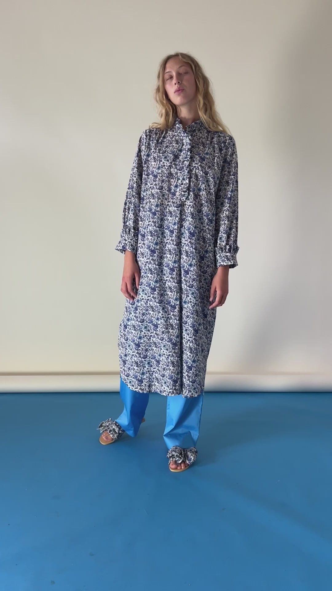 Woman in blue tunic dress with flower pattern, buttons and frills, blue pants and sandals with bows