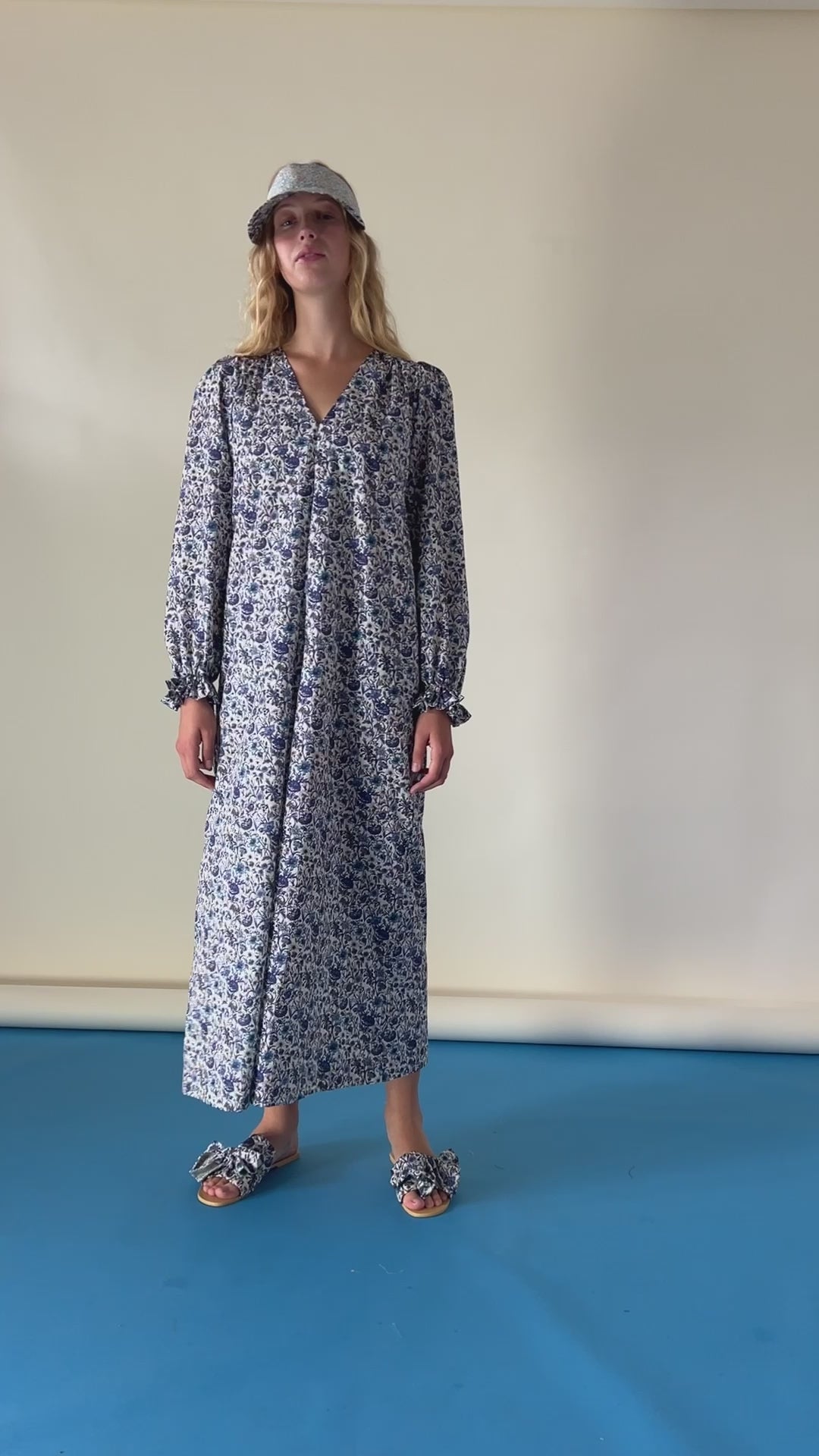 Woman in midi cotton dress with blue flower print, v-neck and ruffled cuffs, sun shade cap and sandals with bows