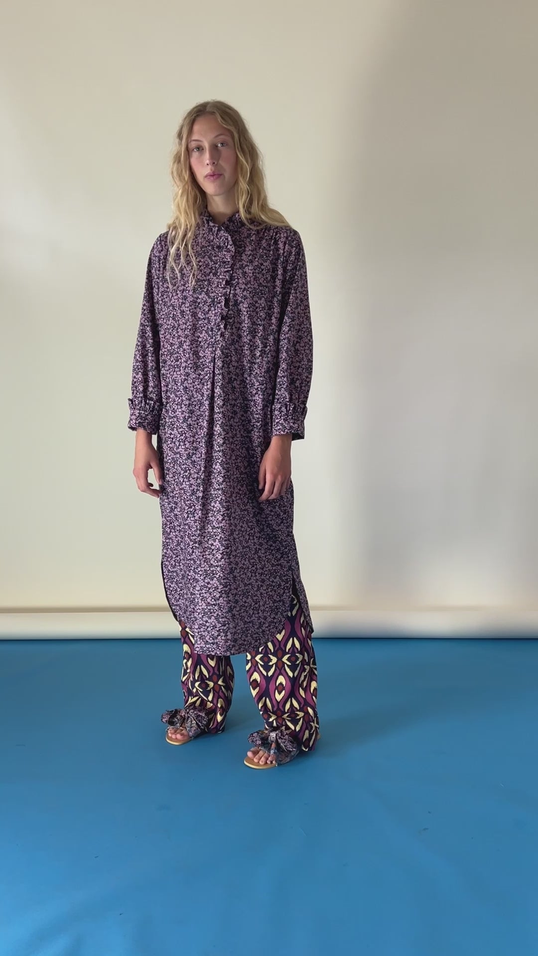 Woman in purple tunic dress with flower pattern, buttons and frills, pants and sandals with bows