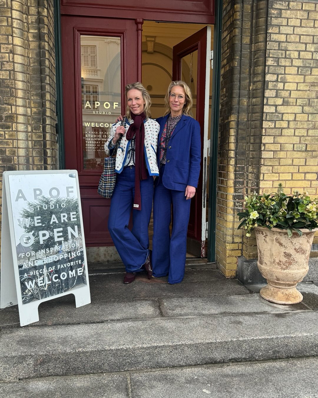 Danish company APOF founders Frederikke and Kathrine Hviid in blue outfits standing outside a brick building with a 'APOF Studio' sign.