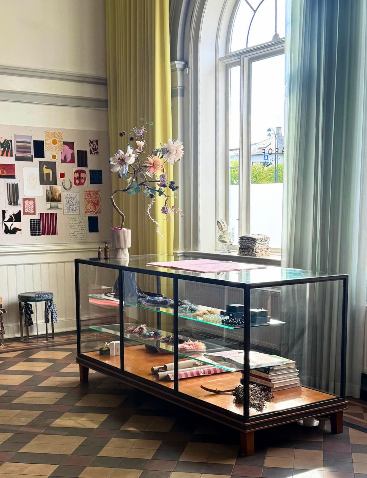 A glass display case with a dark wood base and black metal frame stands in a bright room with patterned tile flooring, large windows, and yellow and blue-gray curtains. The case is styled with books, small objects, and a floral arrangement on top.
