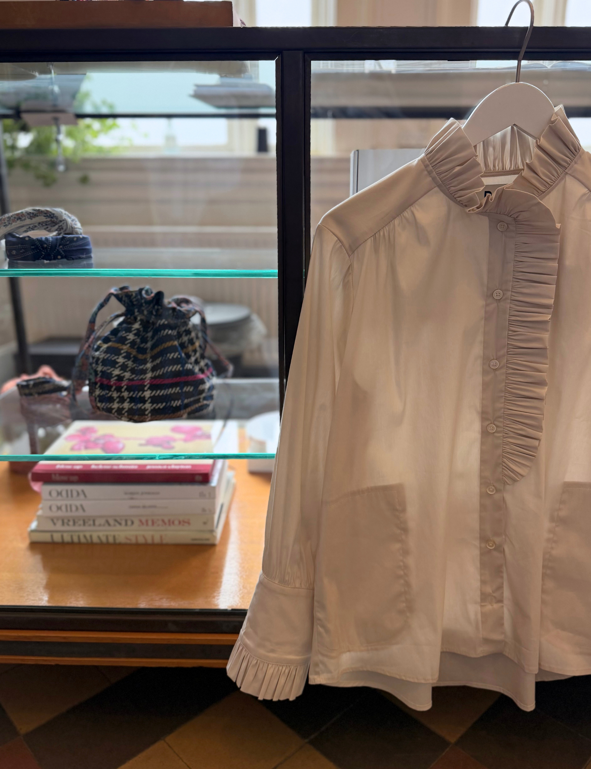 A white long-sleeve blouse with a ruffled high collar and button front, hanging on a hanger in front of a glass display case.