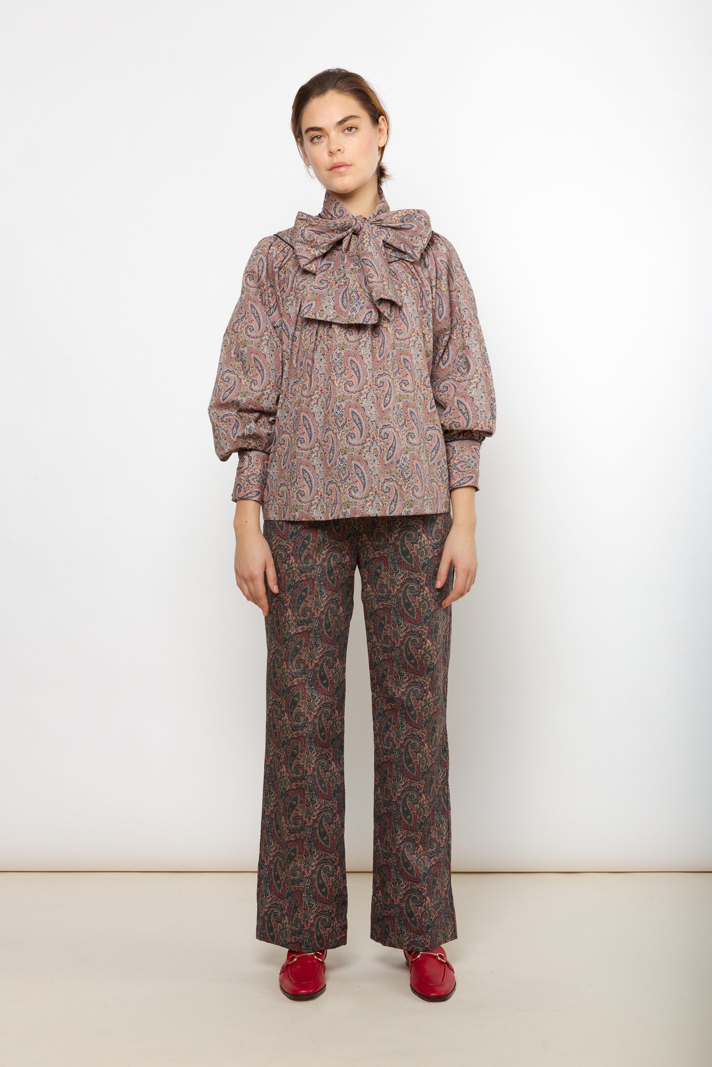Woman in red green and brown paisley patterned cotton pants and long sleeve blouse with large collar bow