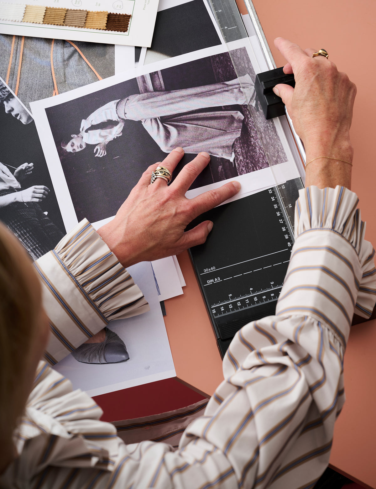 A person in a beige and gray striped blouse works at a desk, placing a black photo negative over printed fashion reference photos and swatches on a peach-colored surface.